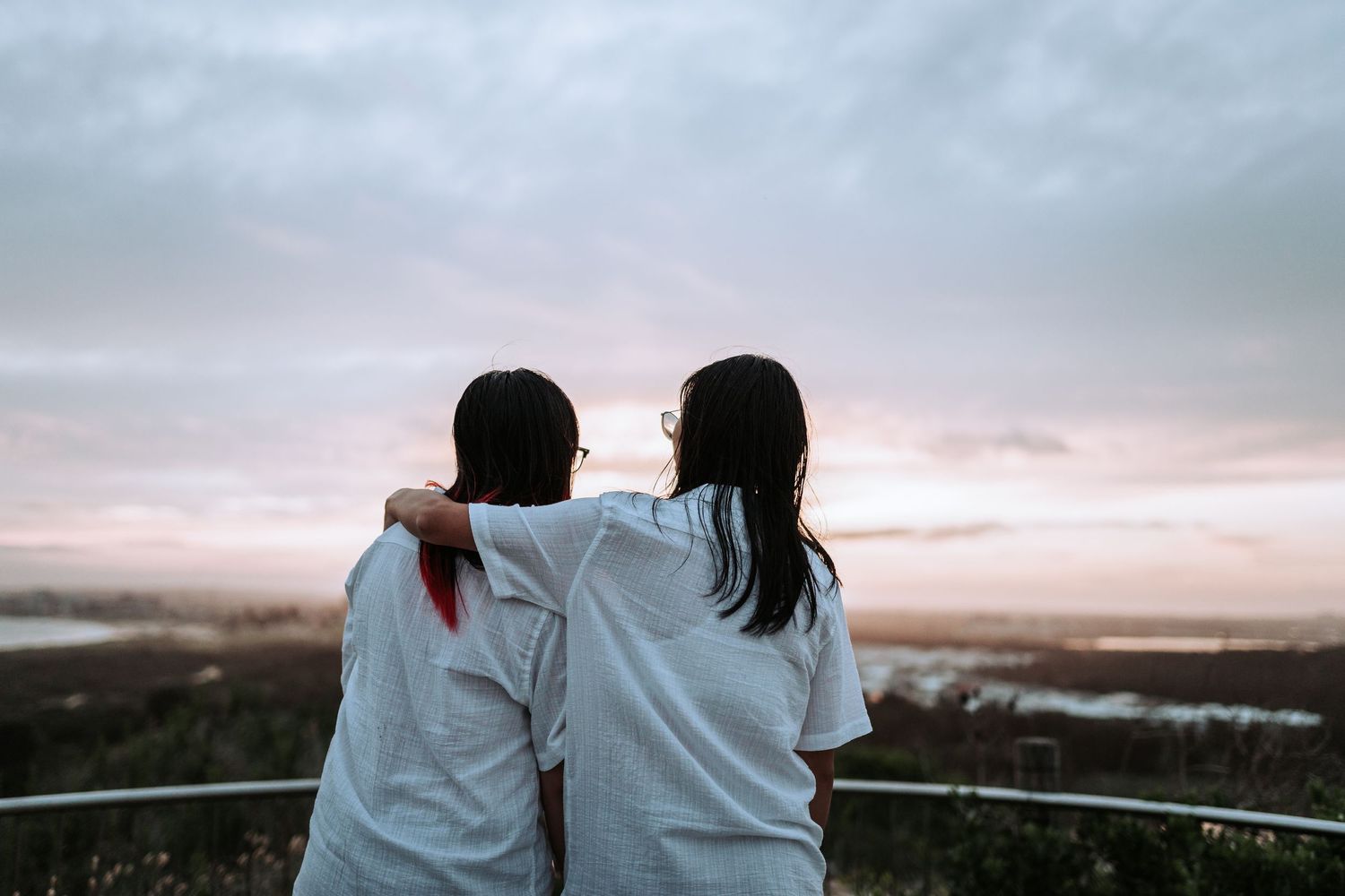 Two people in white shirts sit together watching a sunset over a misty mountain landscape from a viewing point.