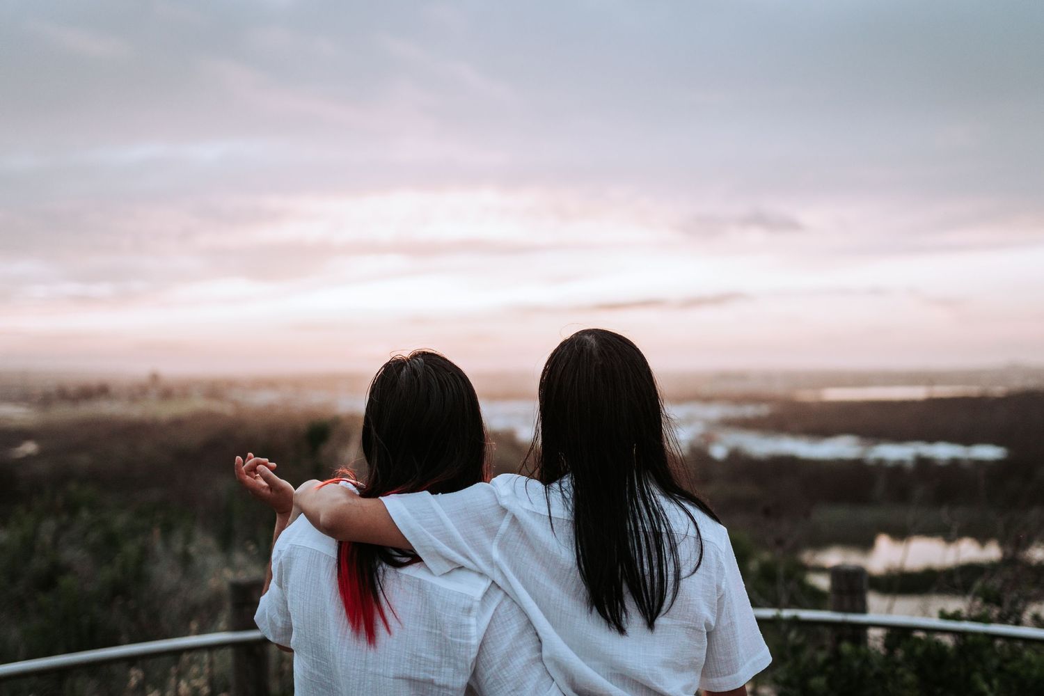 Two people in white shirts watch a misty landscape from an elevated viewpoint during sunset.