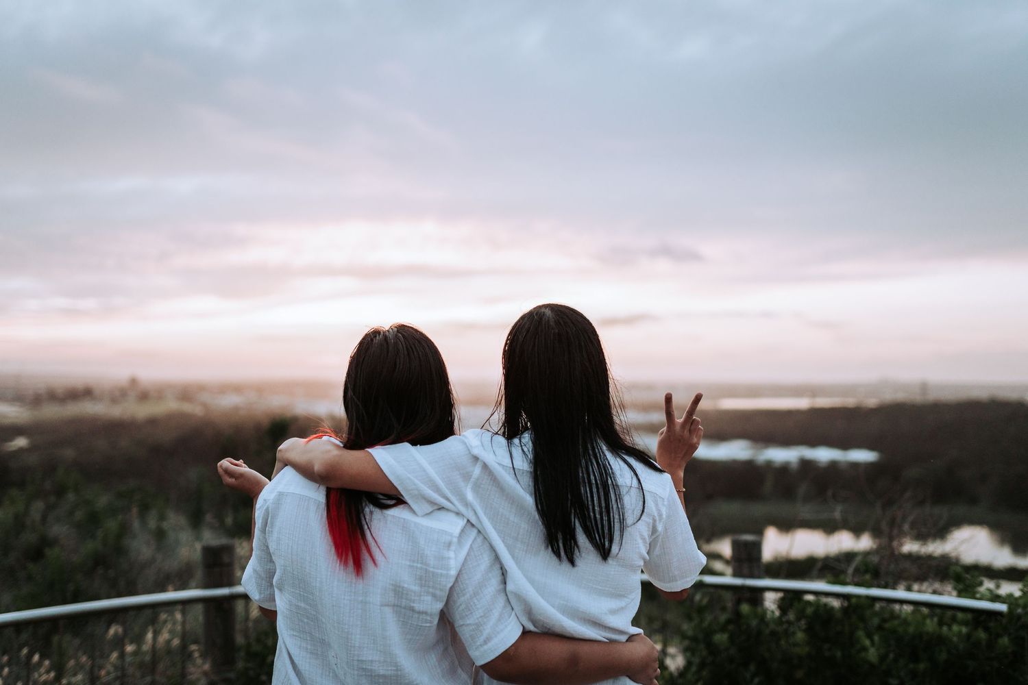 Two friends in white shirts enjoy a sunset view from an elevated viewpoint while making peace signs.