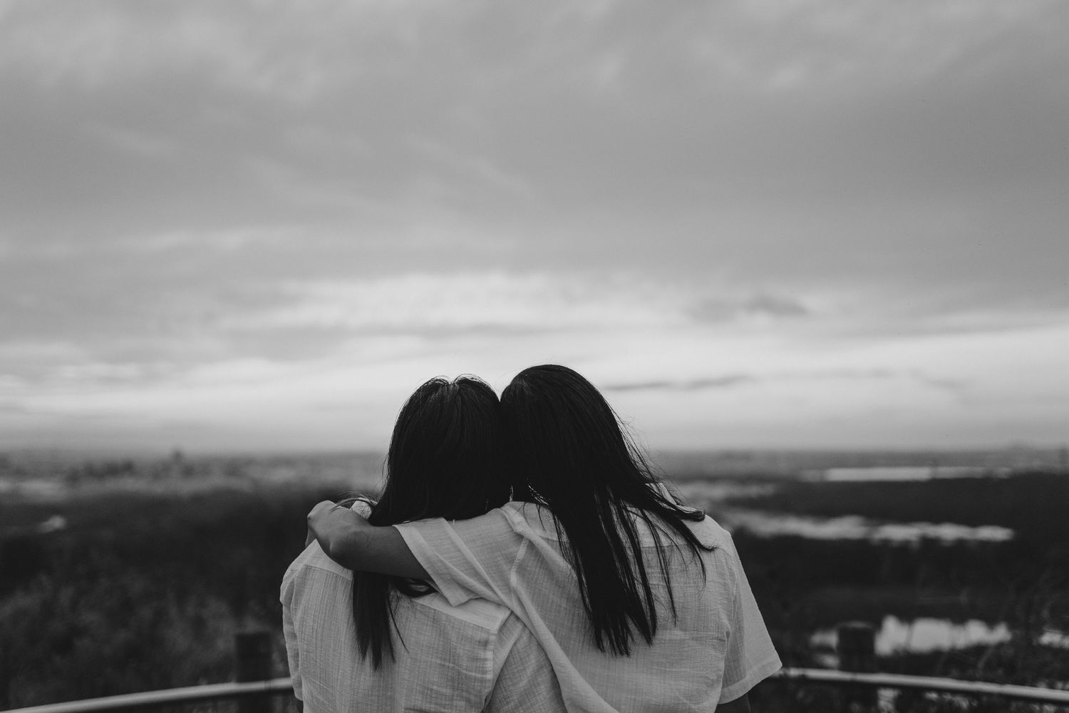 Two figures with long dark hair embrace while looking out over a cloudy landscape in black and white photography.
