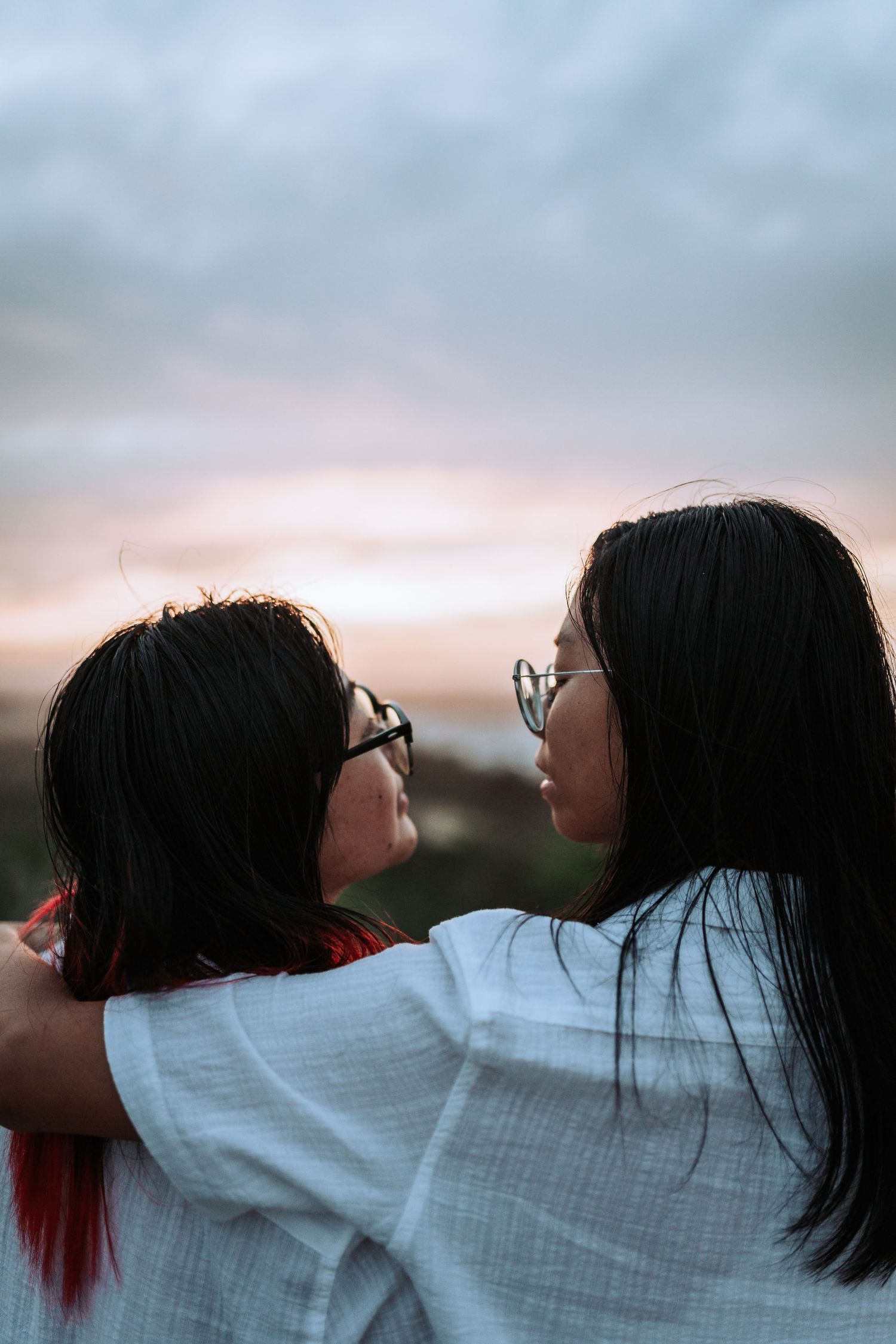 Two friends wearing glasses embrace while watching a sunset with a cloudy sky in the background.
