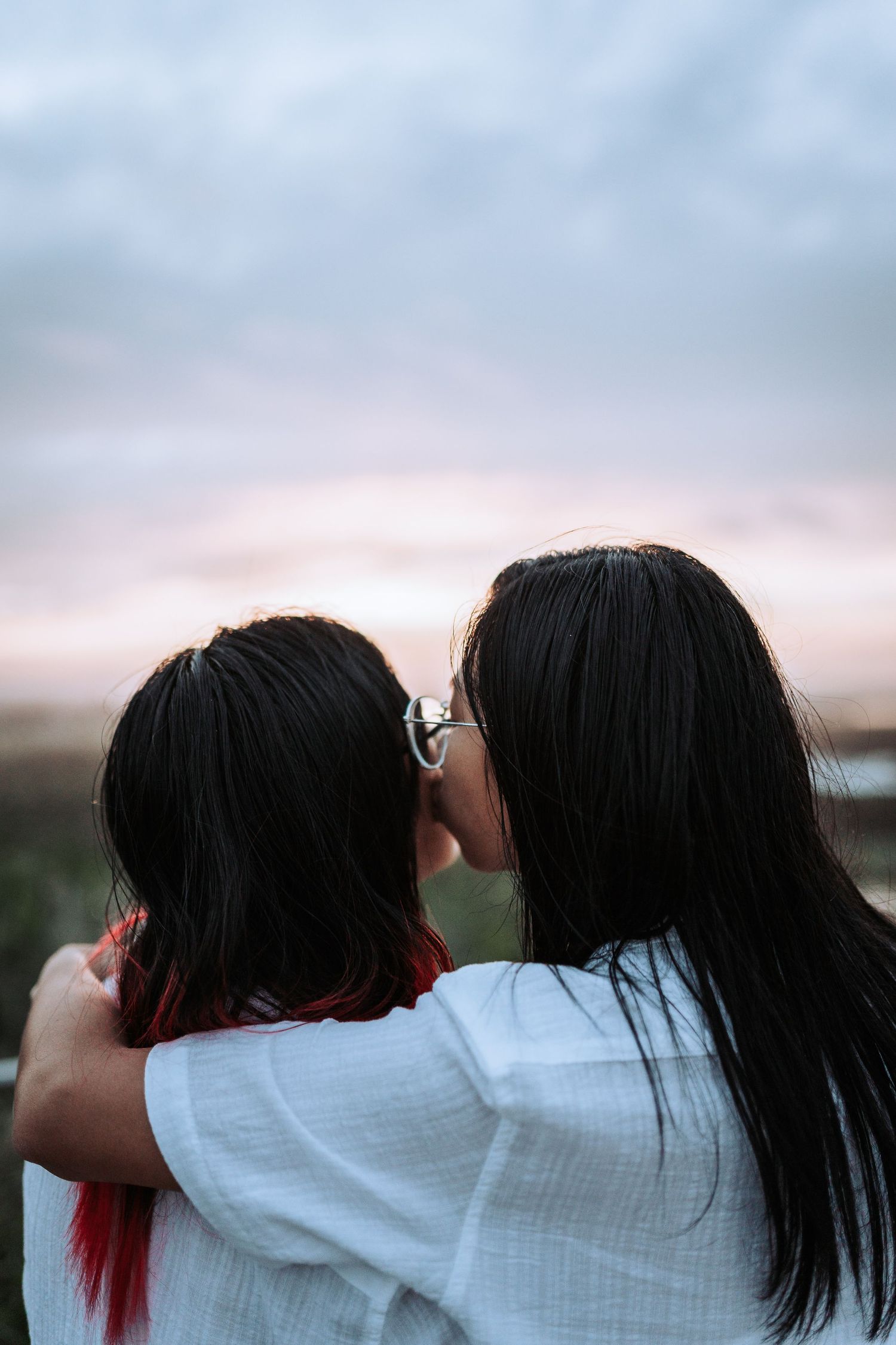 Two figures in white shirts share an intimate moment against a sunset sky with soft clouds.