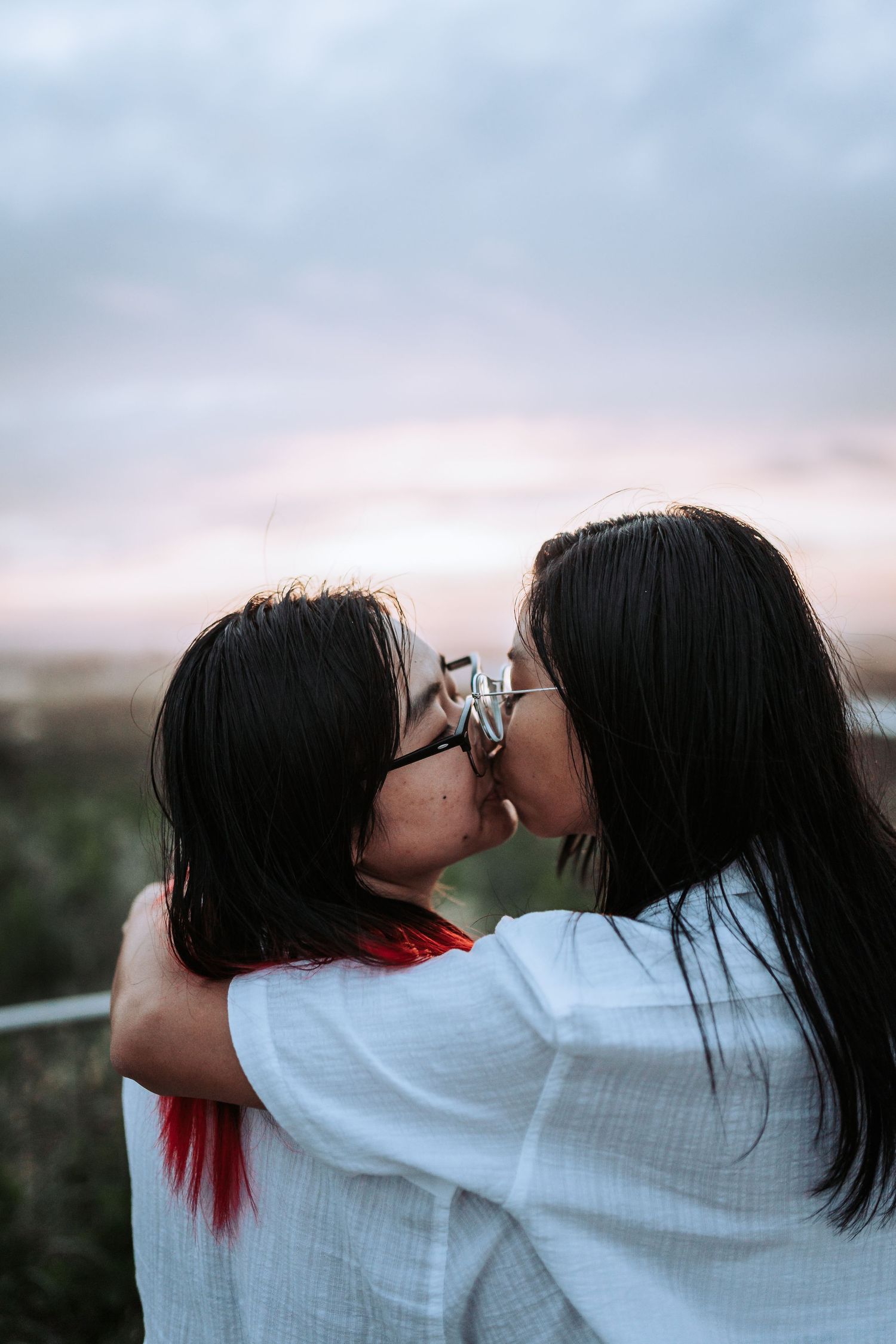 Two people wearing white shirts share a tender embrace against a soft sunset sky outdoors.