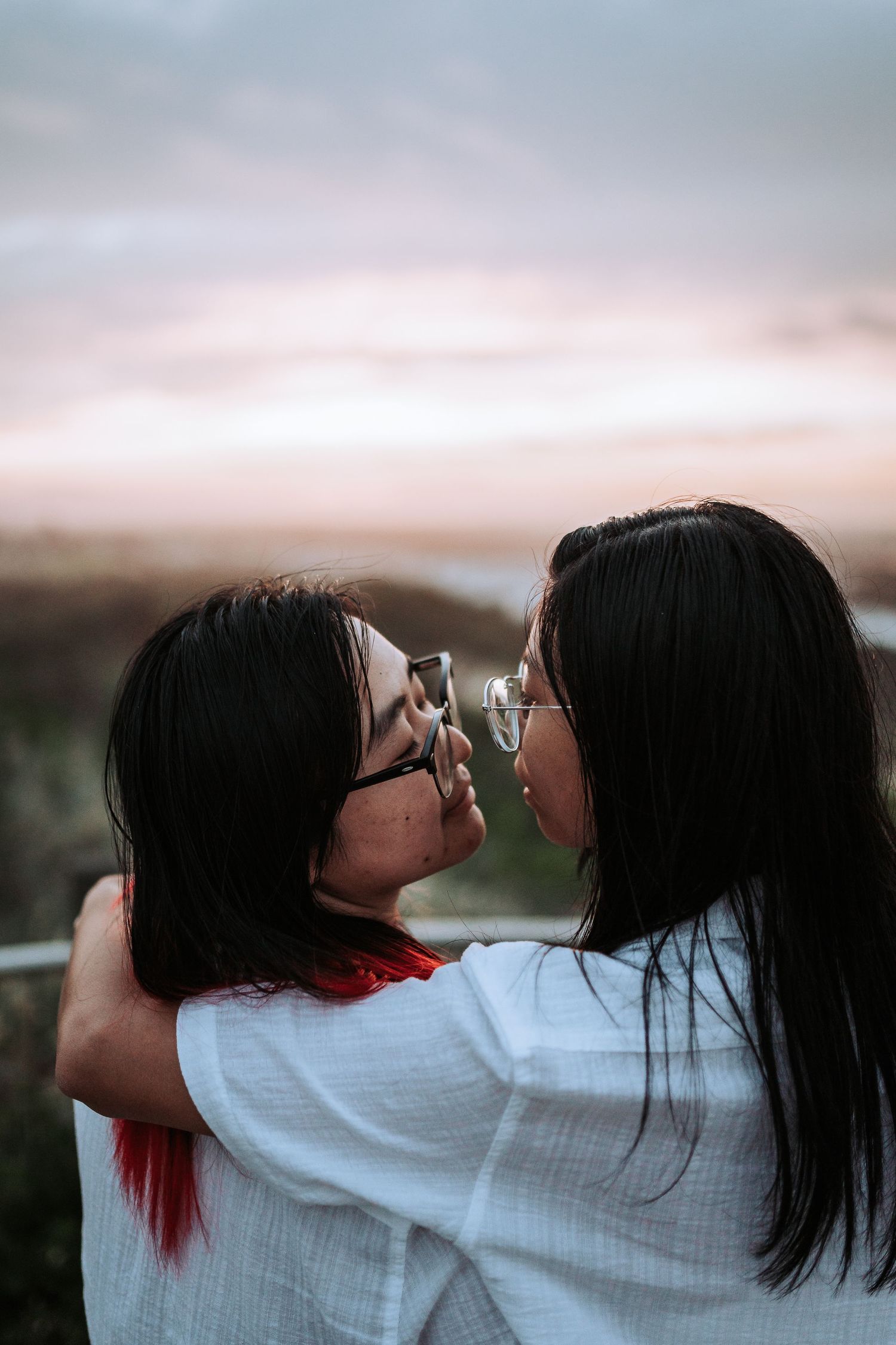 Two people in white shirts share an intimate moment at sunset with one wearing glasses and having red-streaked hair.