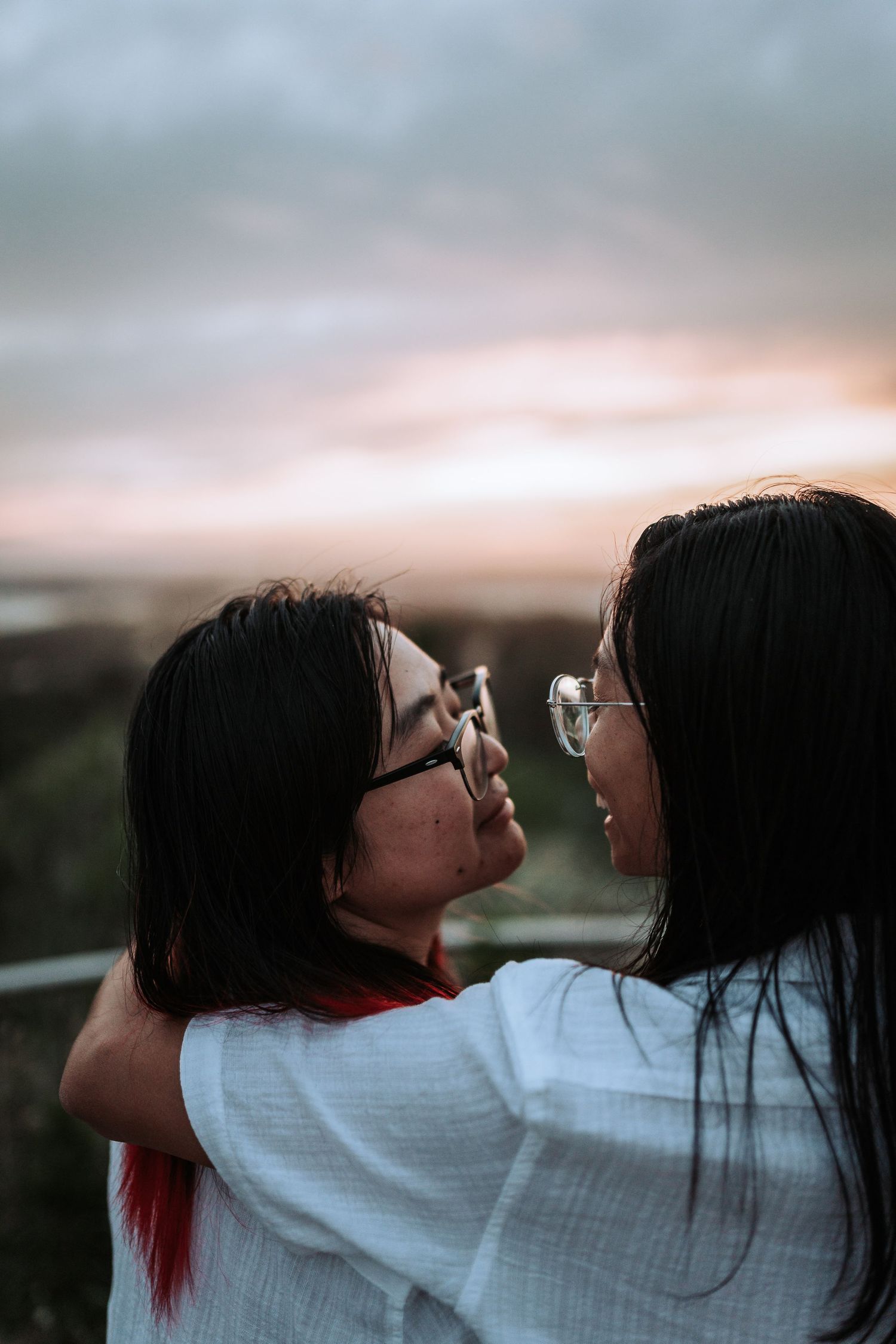 Two people in glasses share an intimate moment against a sunset sky, with one wearing a white shirt.