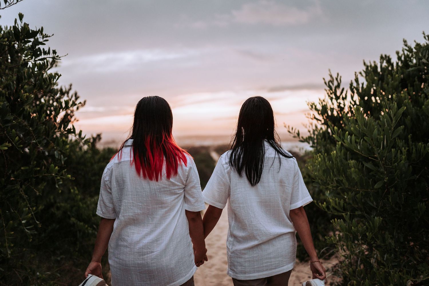 Two people in white shirts with long hair holding hands while watching sunset between trees on a cloudy evening.