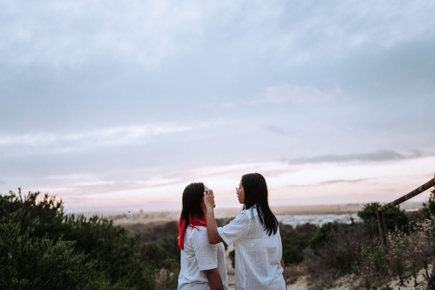 Two figures in white clothing stand together against a cloudy sunset sky on a hilltop with desert brush.