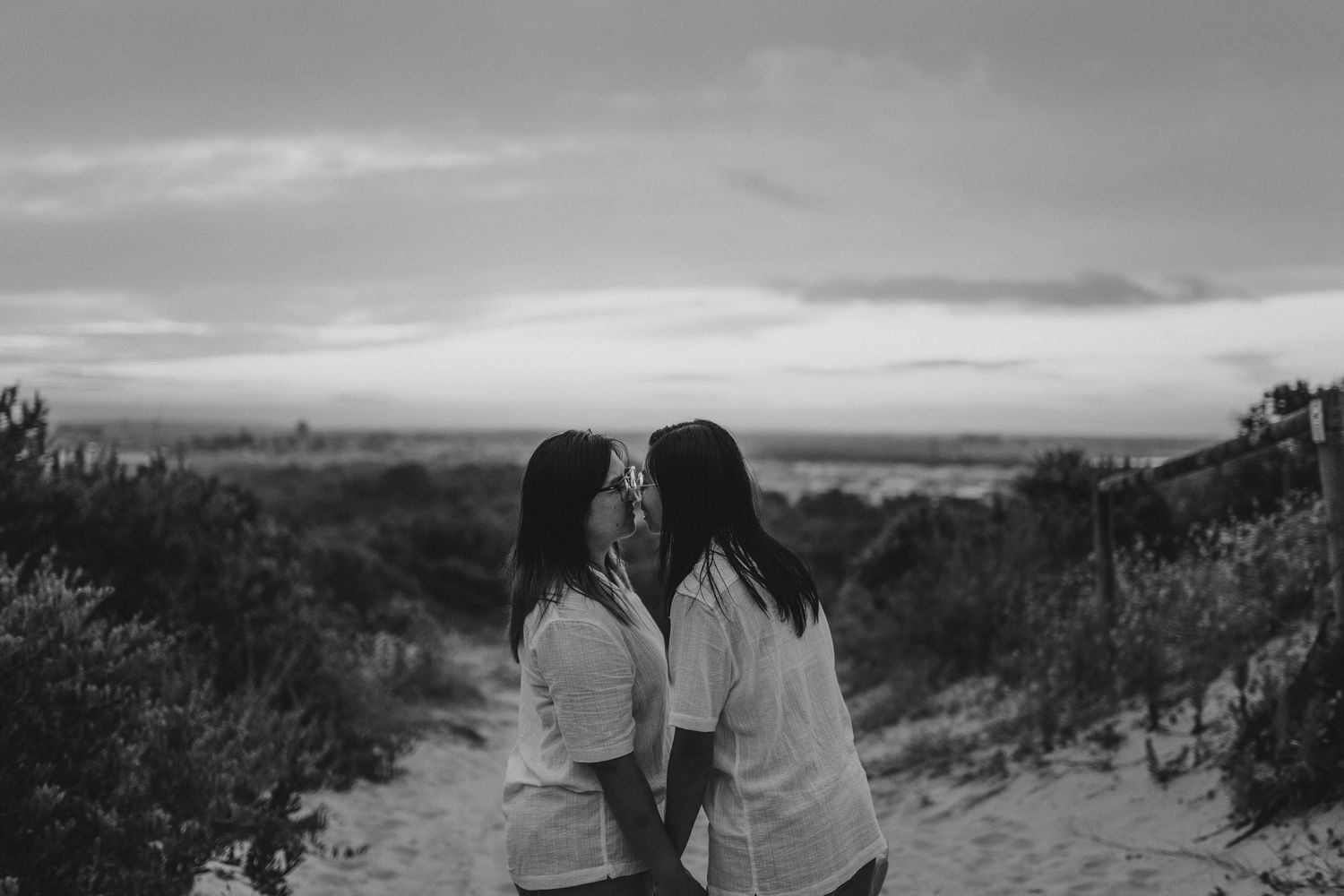A black and white scenic landscape photograph showing two people sitting close together on a hilltop at sunset.