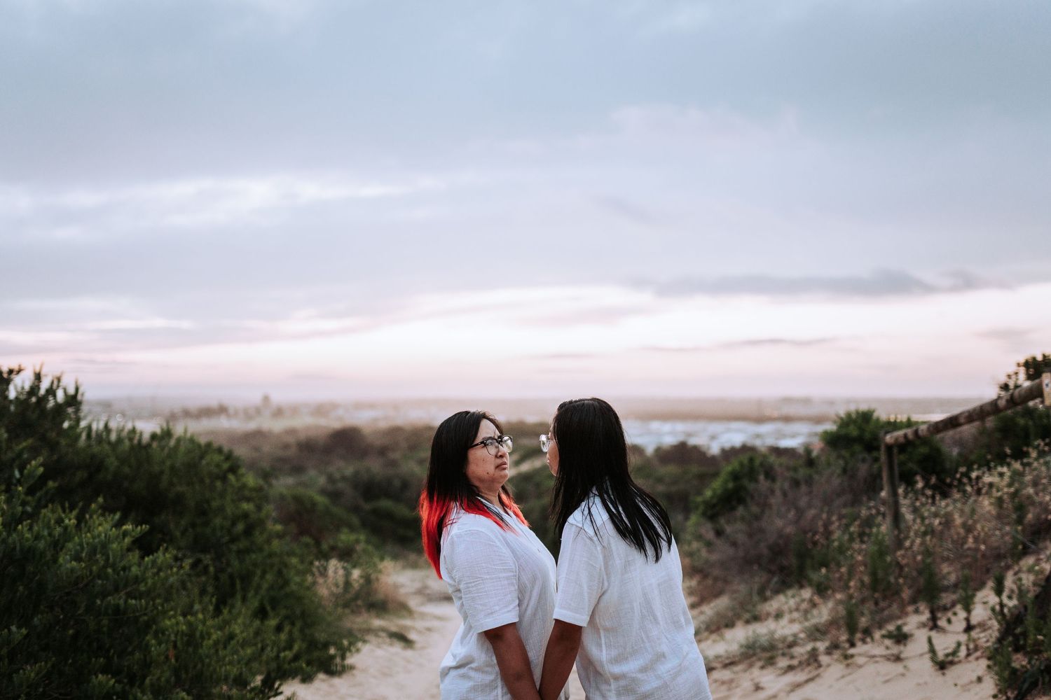 Two people wearing white tops share a tender moment on a hilltop at sunset with coastal scenery in the background.