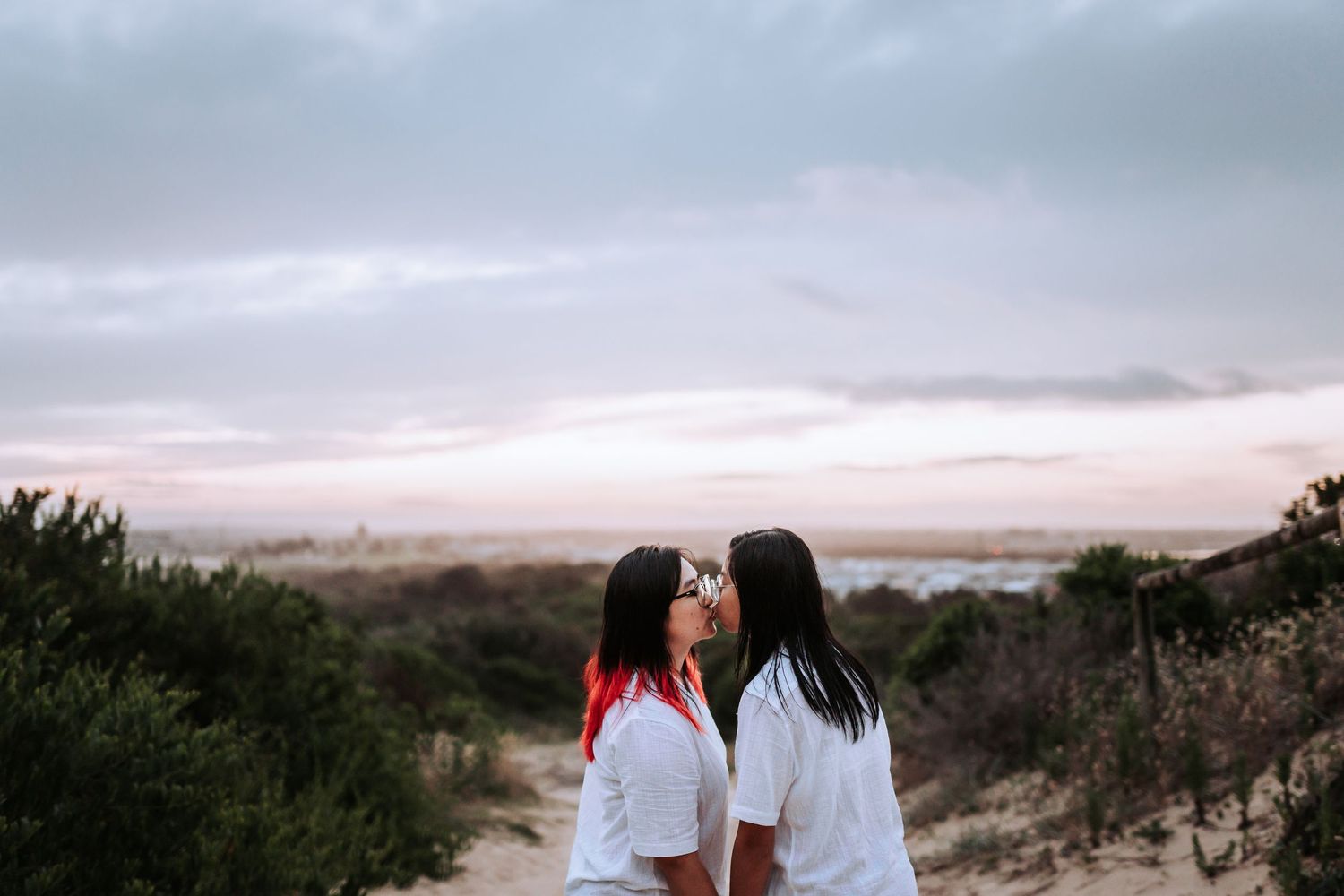 Two people in white shirts stand together on a hilltop path at sunset with moody cloudy sky in background.