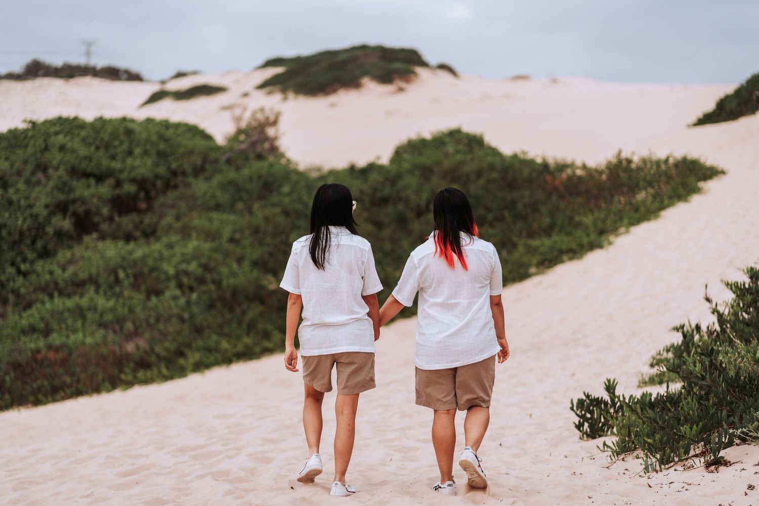 Two people in white shirts and shorts walk together on a sandy path bordered by green shrubs and dunes.