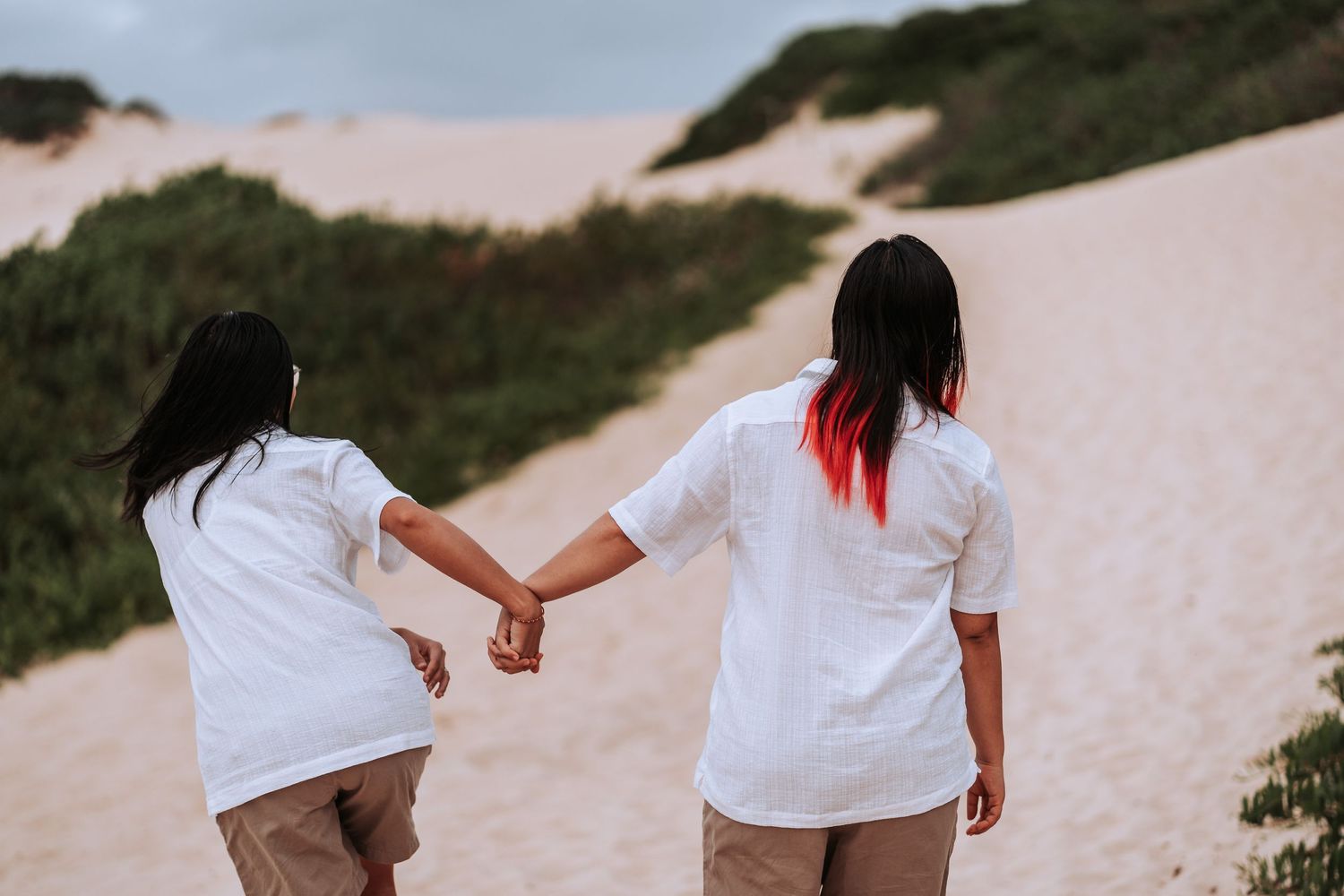 Two people wearing white shirts and beige pants hold hands while walking on sandy dunes at sunset.