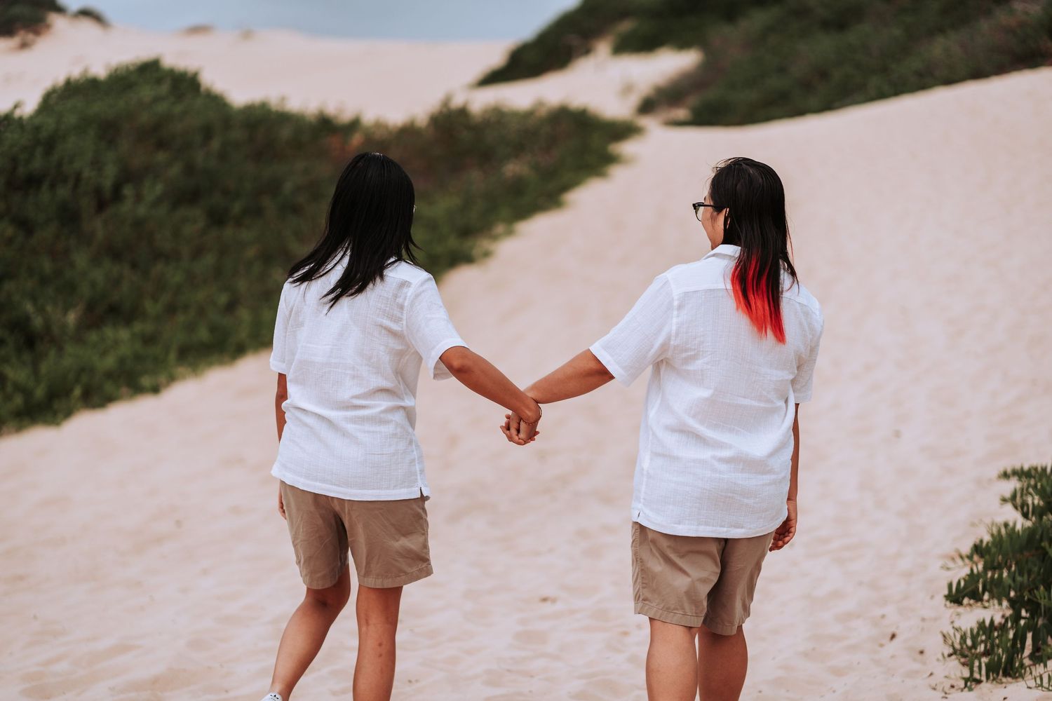 Two people in white shirts and shorts hold hands while walking on a sandy coastal path at sunset.