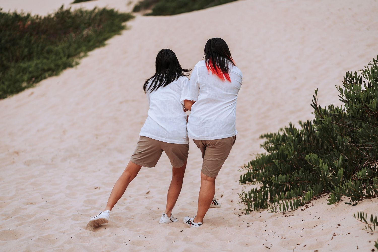 Two people in white tops and beige shorts walk together on a sandy path bordered by green shrubs.