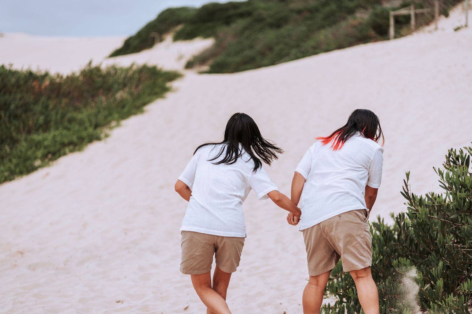 Two people in white shirts and khaki shorts run up a sandy coastal path together on a sunny day.
