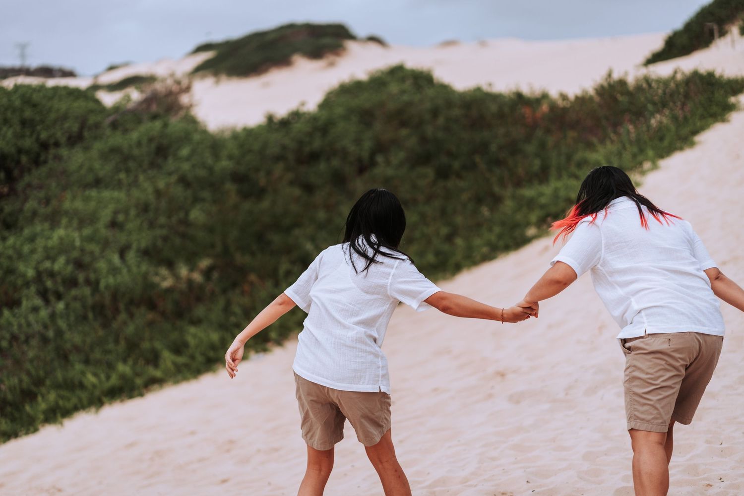 Two people in white shirts and khaki shorts walking hand in hand across a sandy path with green vegetation.