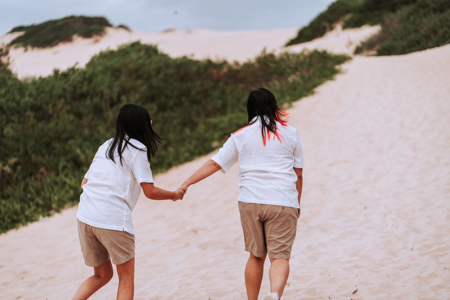 Two people in white shirts and khaki shorts hold hands while climbing sandy dunes together.