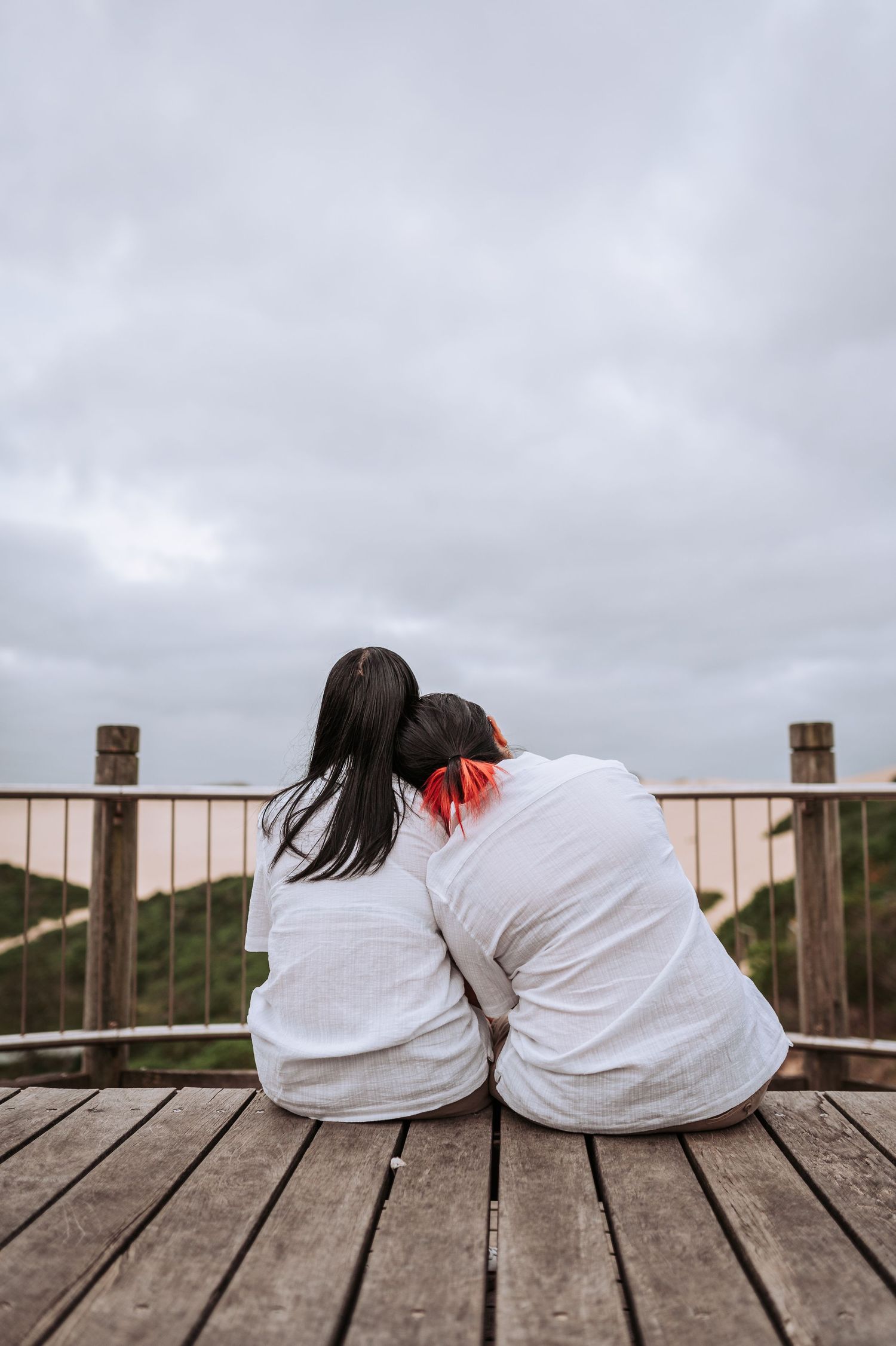 Two people in white shirts sit close together on a wooden deck overlooking a cloudy landscape.