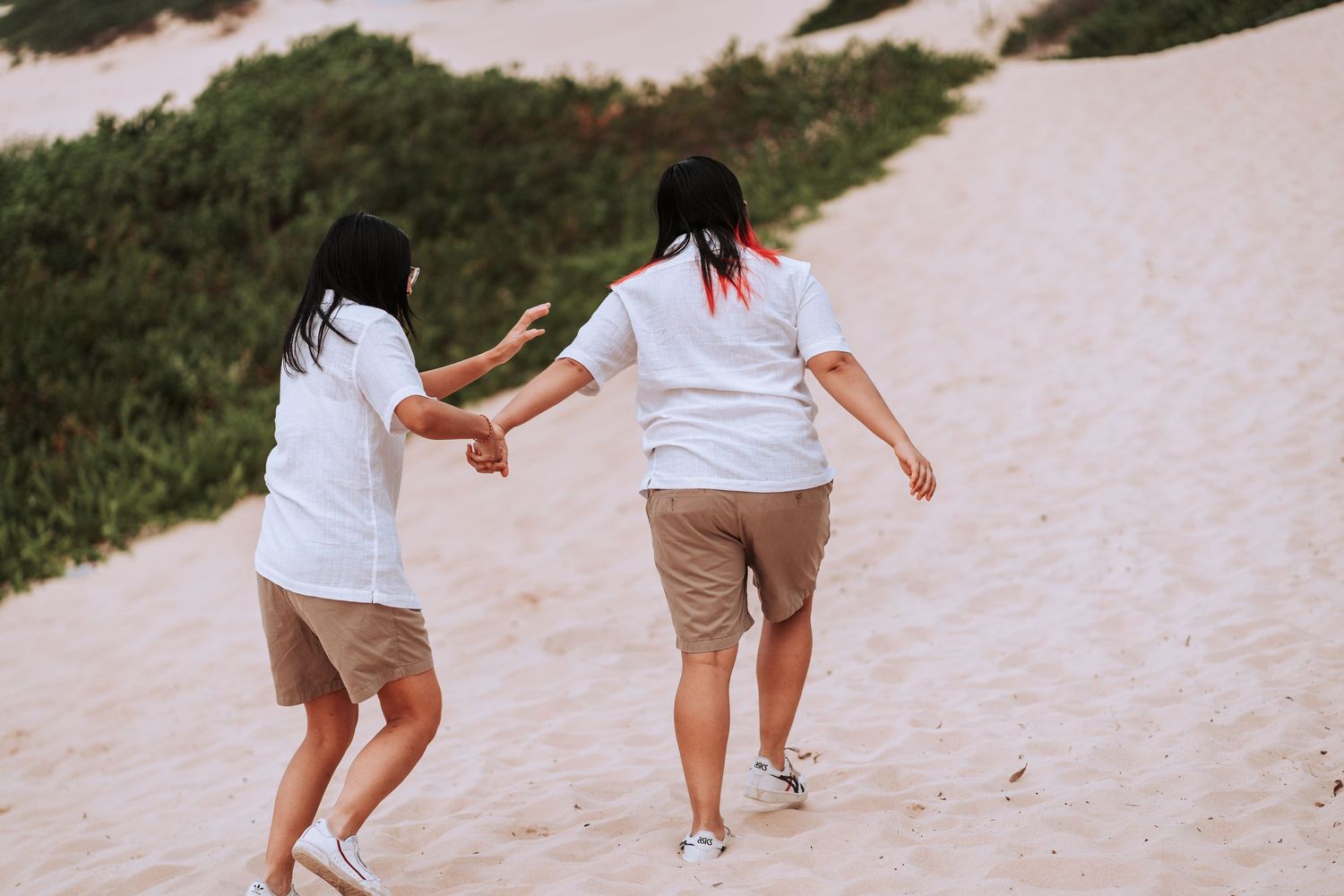 Two people in white shirts and khaki shorts hold hands while walking up a sandy path together.