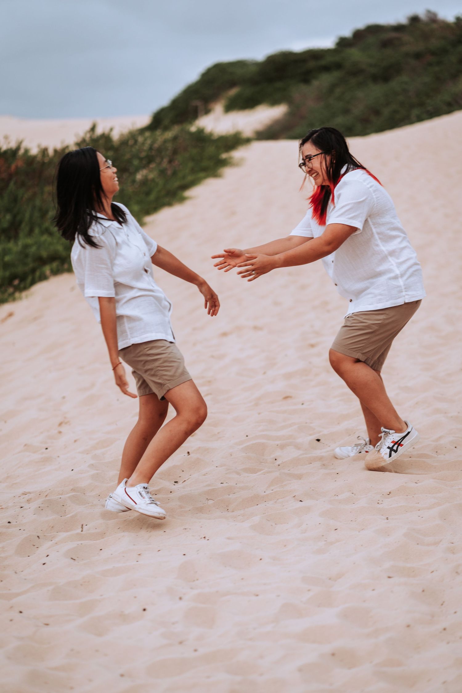 Two people in white shirts and khaki shorts dancing and laughing together on a sandy beach during sunset.