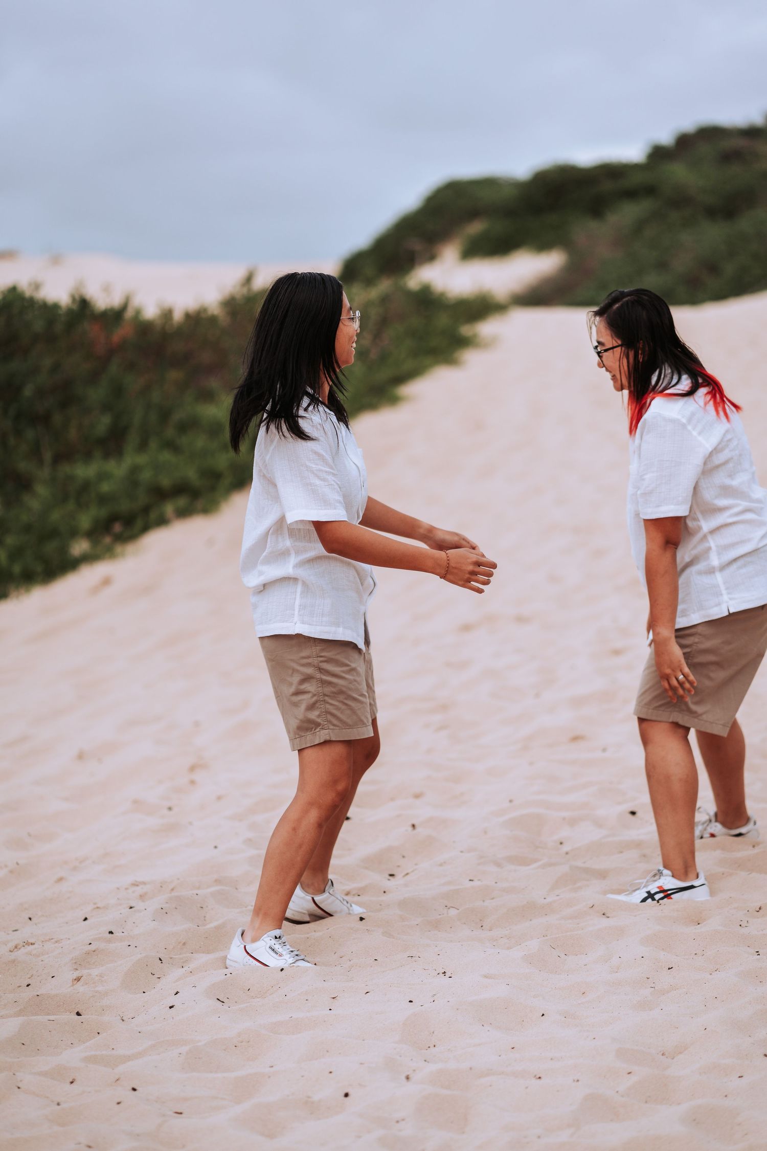 Two people in matching white shirts and khaki shorts playing together on a sandy beach slope during sunset.