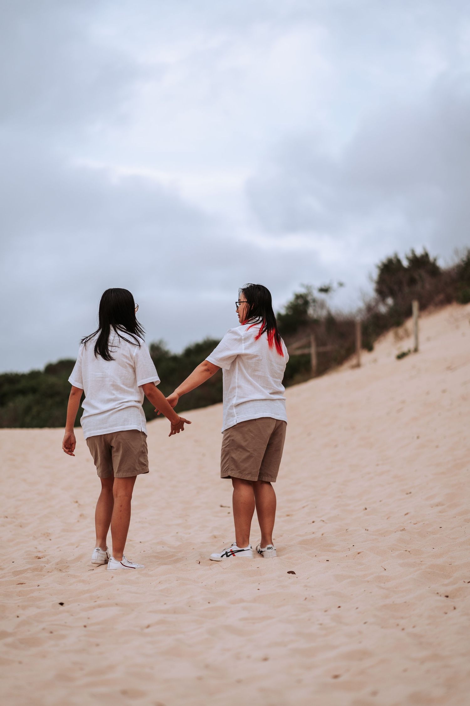 Two people in white shirts and khaki shorts holding hands while walking on a sandy beach slope under cloudy skies.