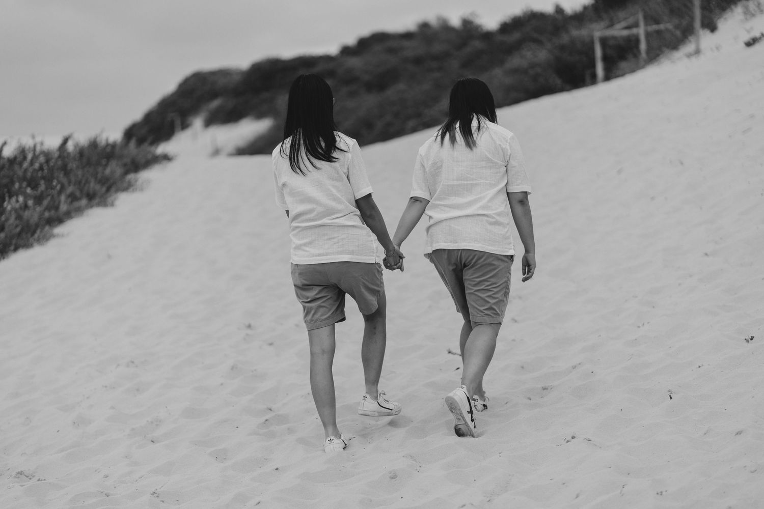 Two people in matching white shirts and shorts walk together holding hands on a sandy beach slope in black and white.