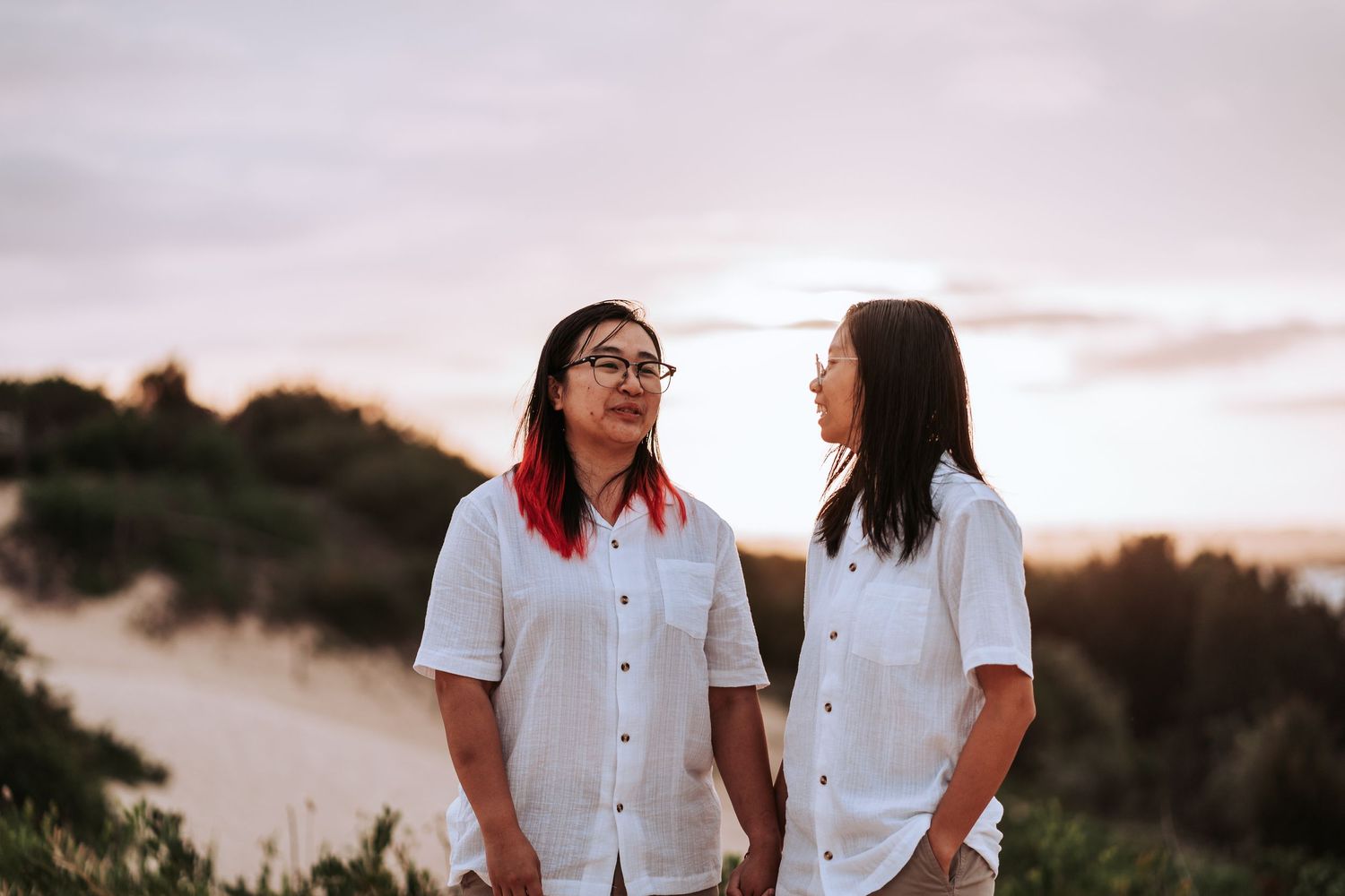 Two people in white button-up shirts smile at each other against a sunset backdrop near the ocean.