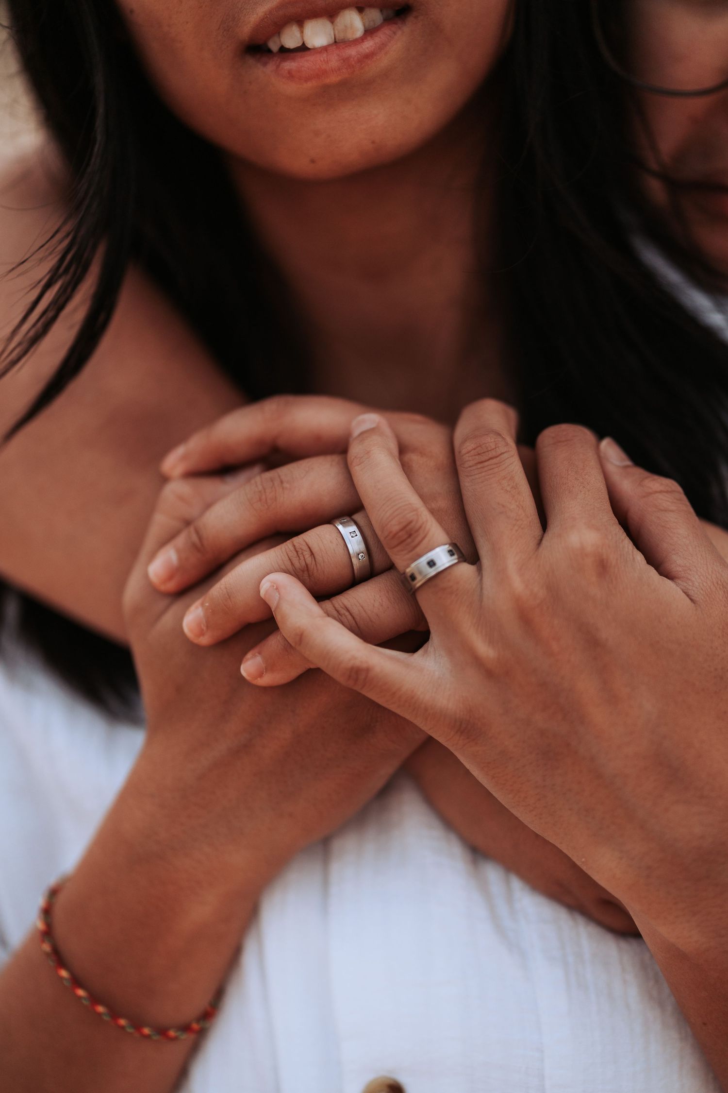 Close-up of hands wearing wedding bands with a white dress and beaded bracelet visible in the background.