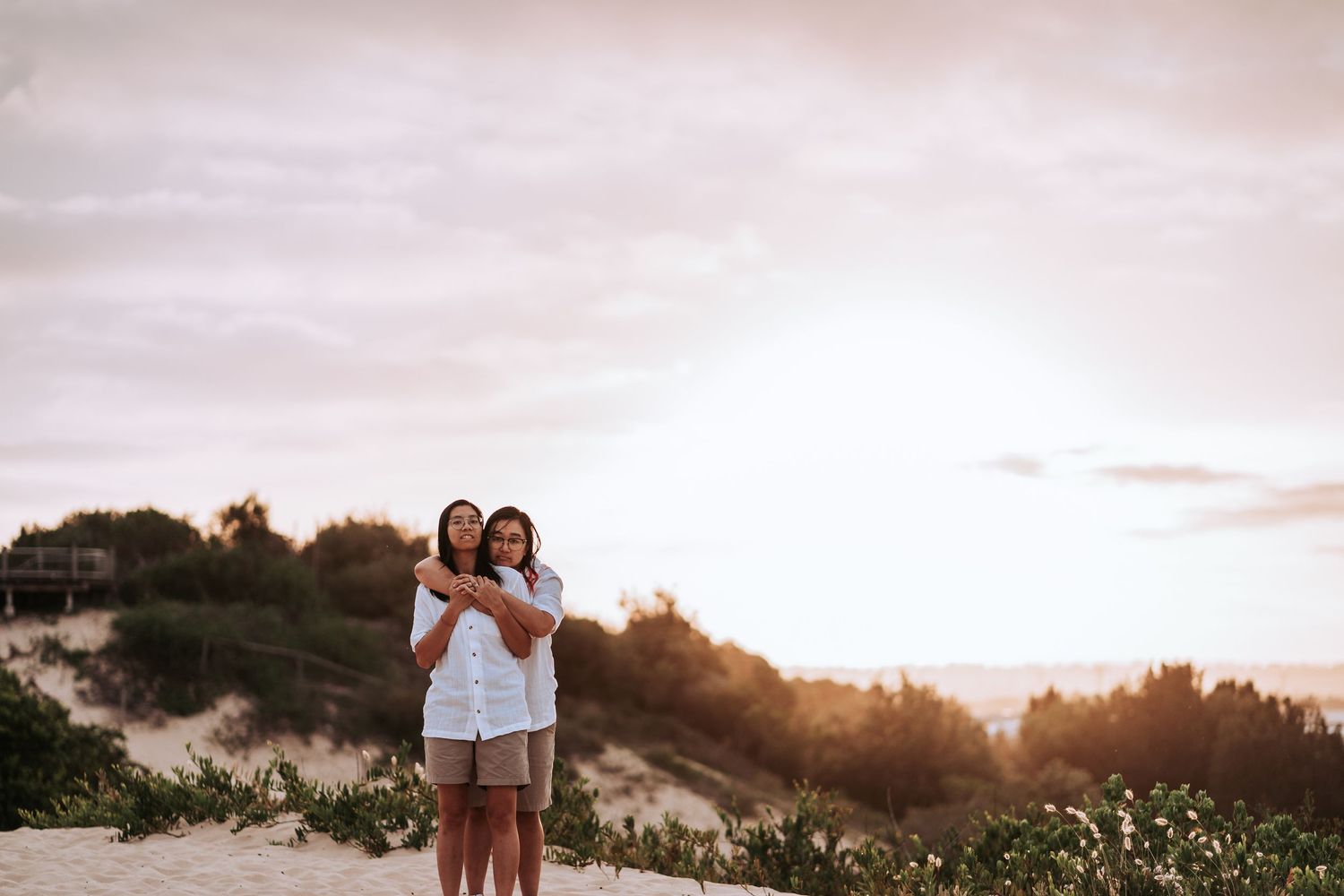 Two people standing together on a hilltop against a sunset sky with vegetation in the foreground.