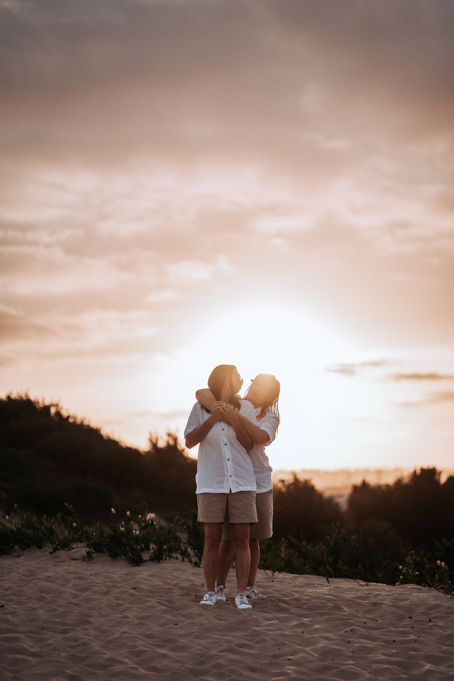 Couple embraces on sandy beach during golden sunset with silhouetted trees in background.