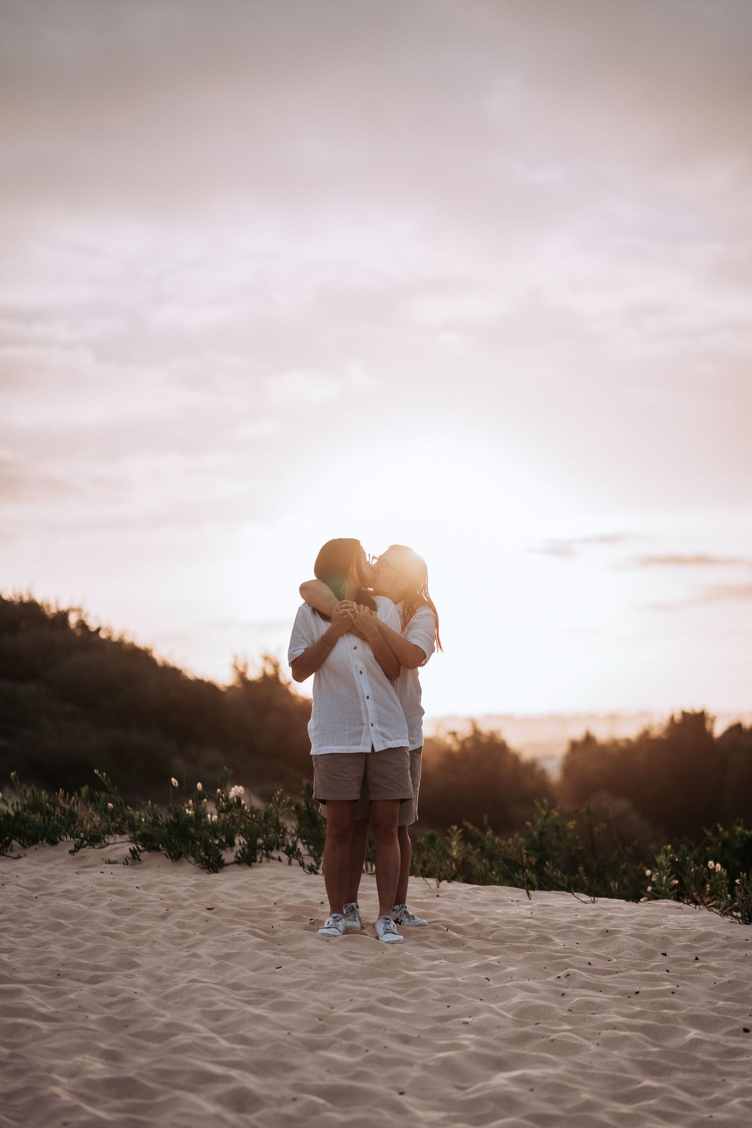 A couple embraces on a sandy hill at sunset with a glowing pink and orange sky in the background.