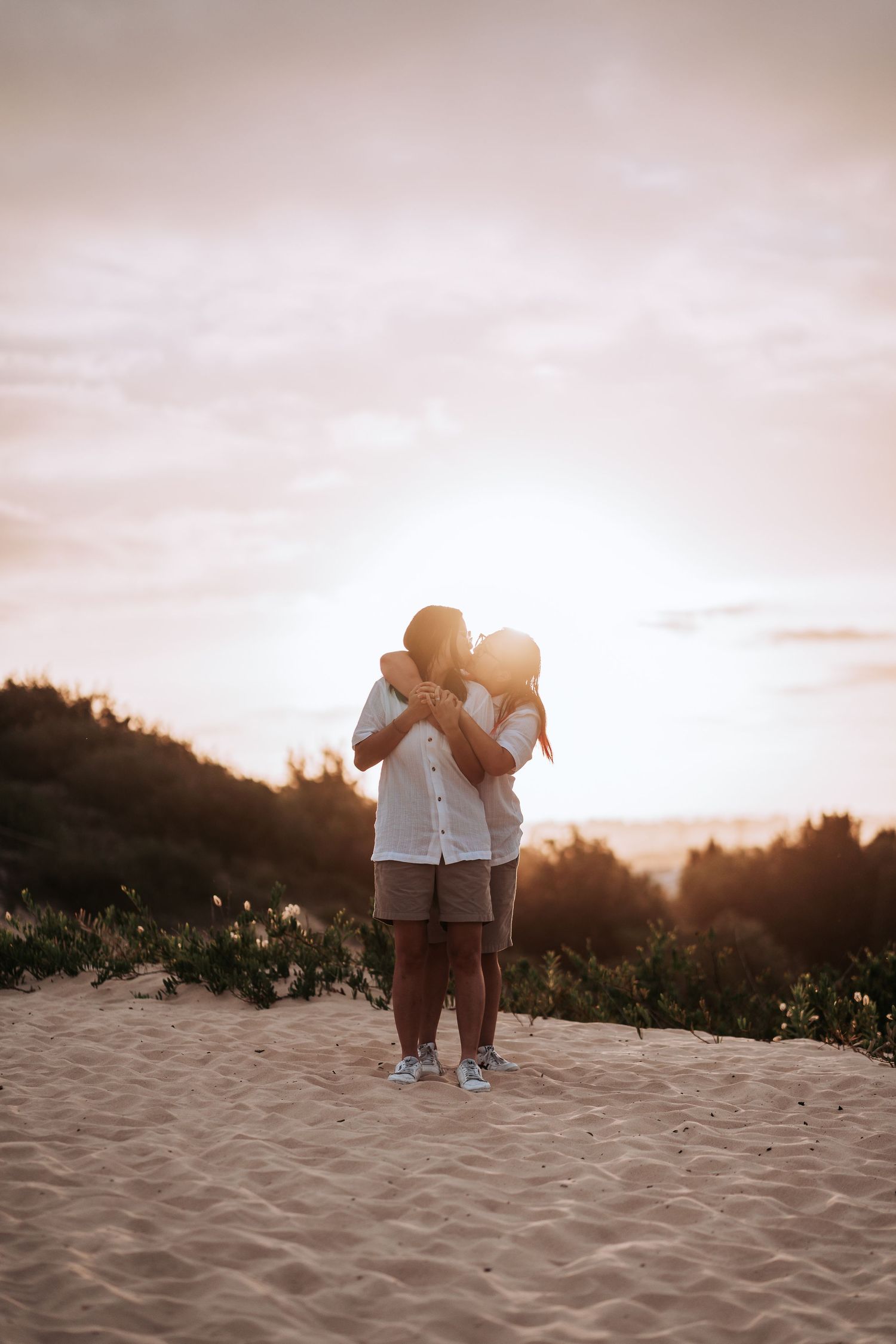 A couple embraces on a sandy beach during sunset with silhouetted trees in the background.
