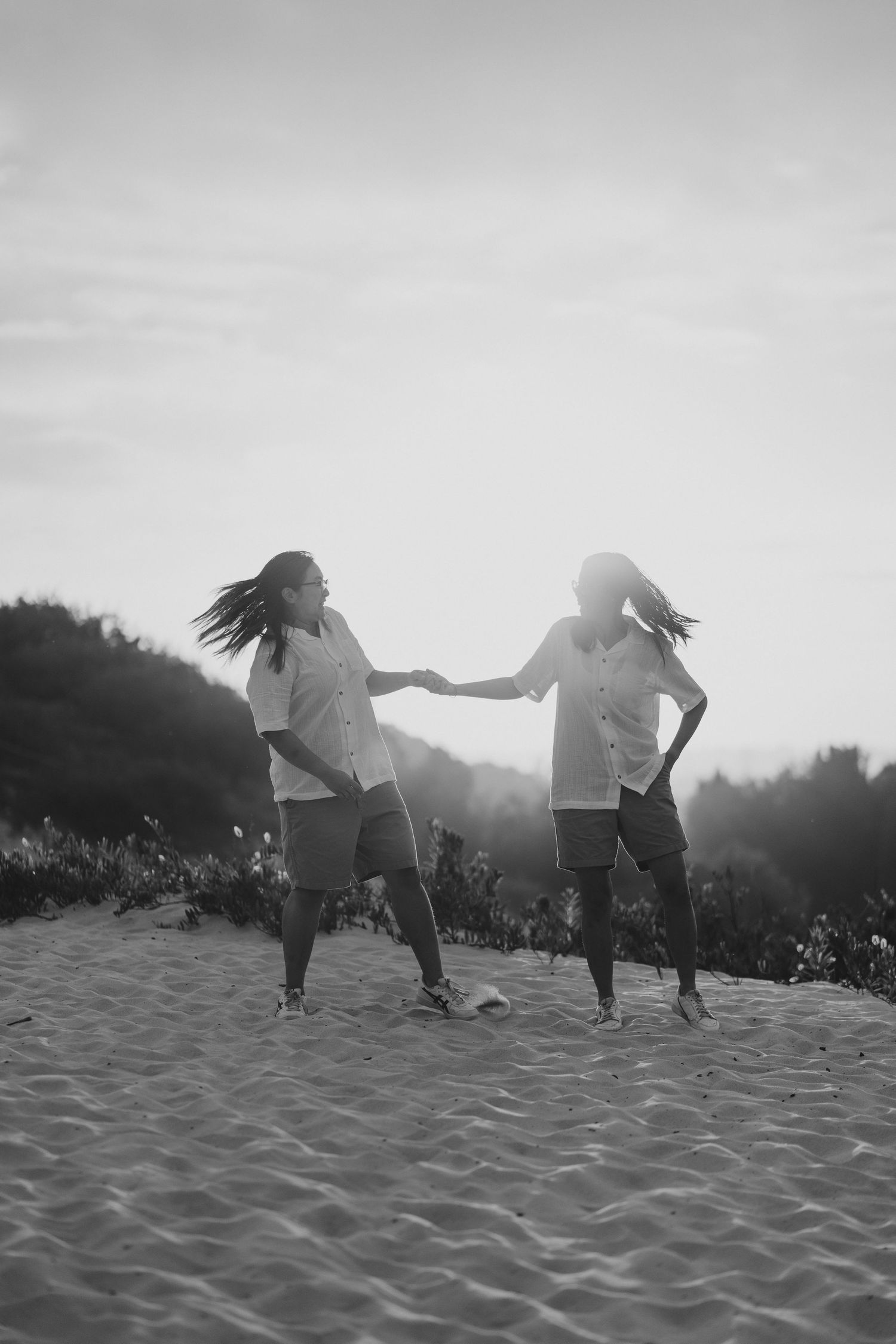 Two figures dancing together on sandy beach at sunset, silhouetted against bright sky in black and white photo.