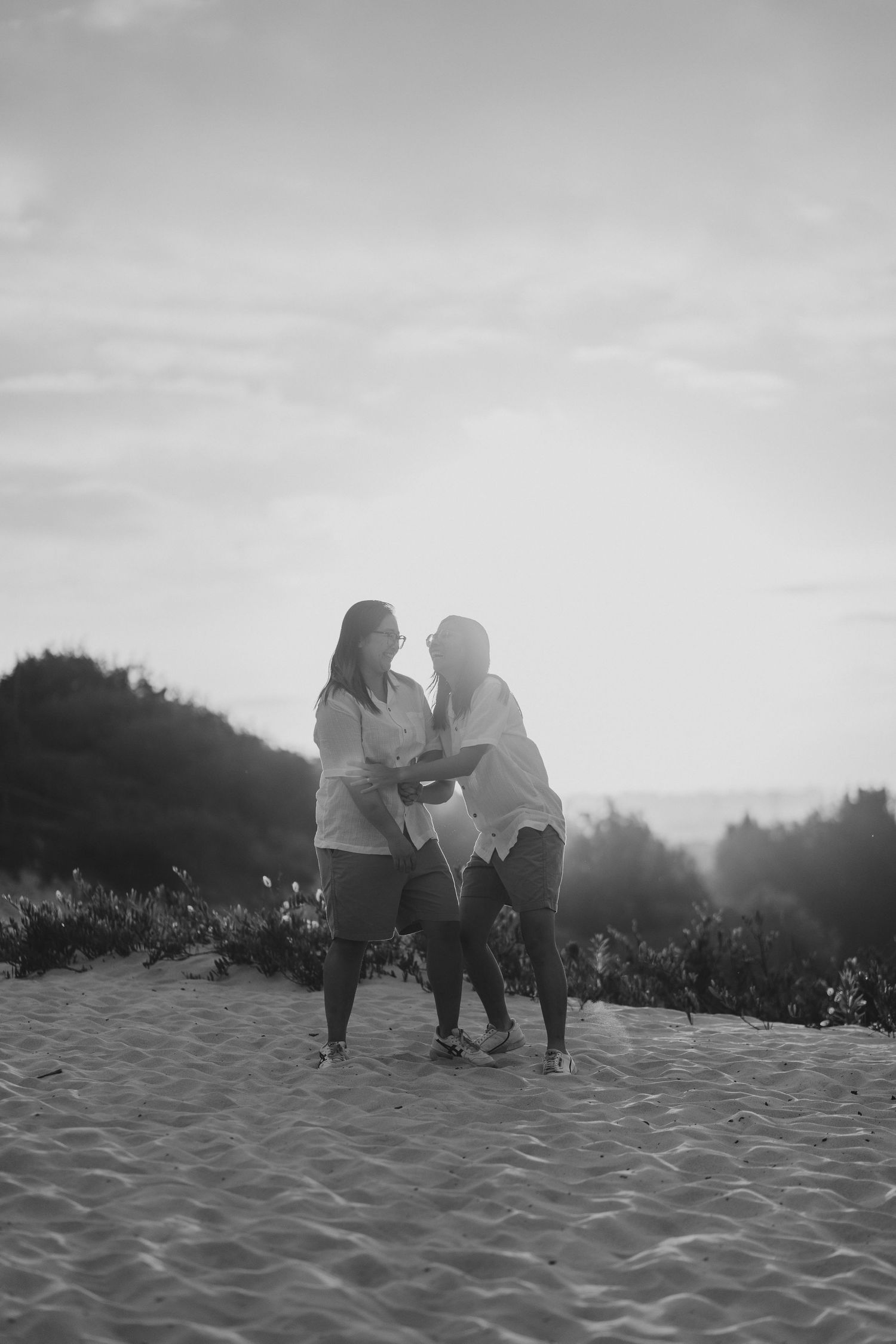 Two figures stand together on sandy dunes against misty mountains in a dreamy black and white photo.