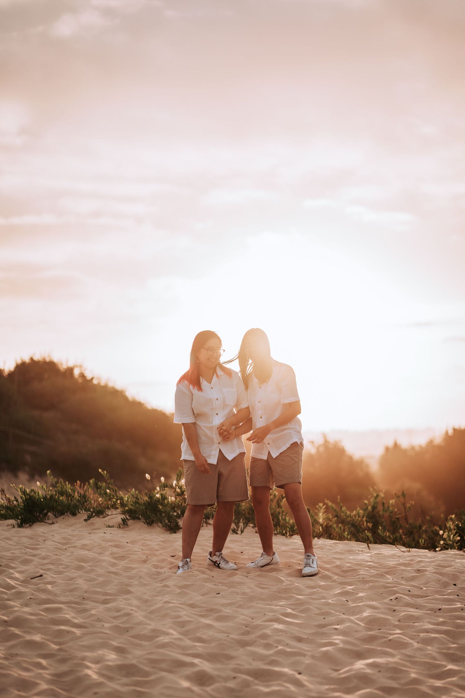 Couple in casual white shirts laughing together on sandy beach during golden sunset.