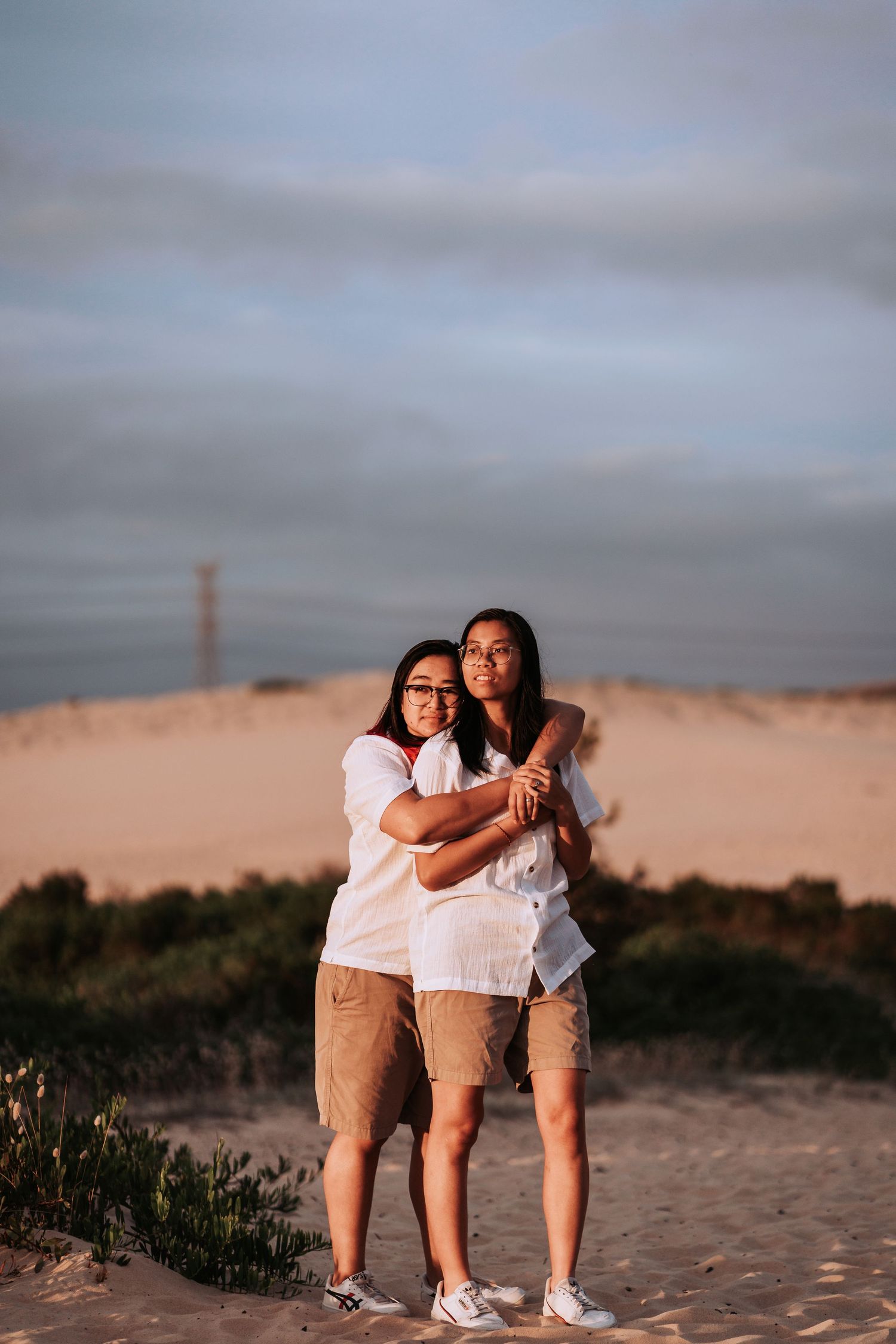 Two people in white shirts and tan shorts embrace while standing on sand dunes at sunset.