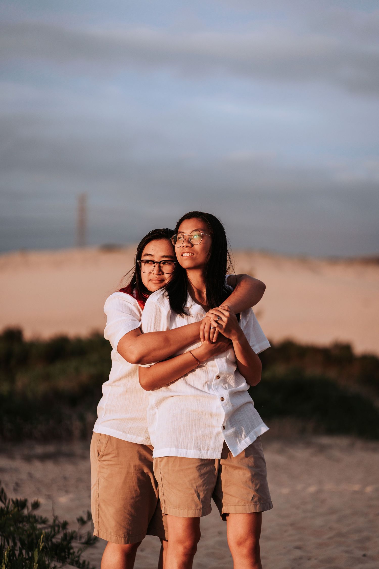 A couple in matching white tops and khaki shorts embrace while standing on a sandy beach at sunset.