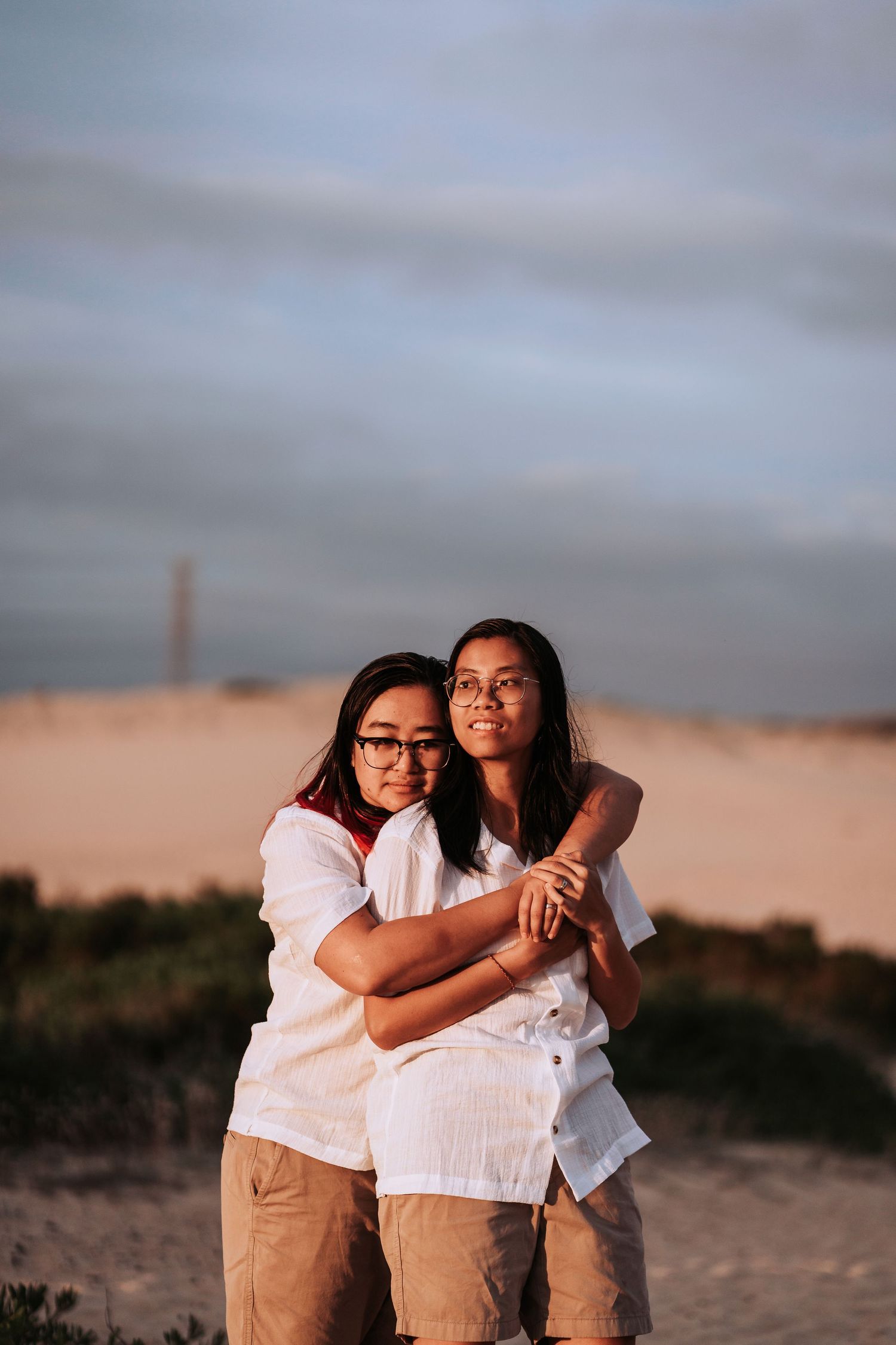 Two people in white shirts and khaki pants embrace while standing outdoors at sunset against a cloudy sky.