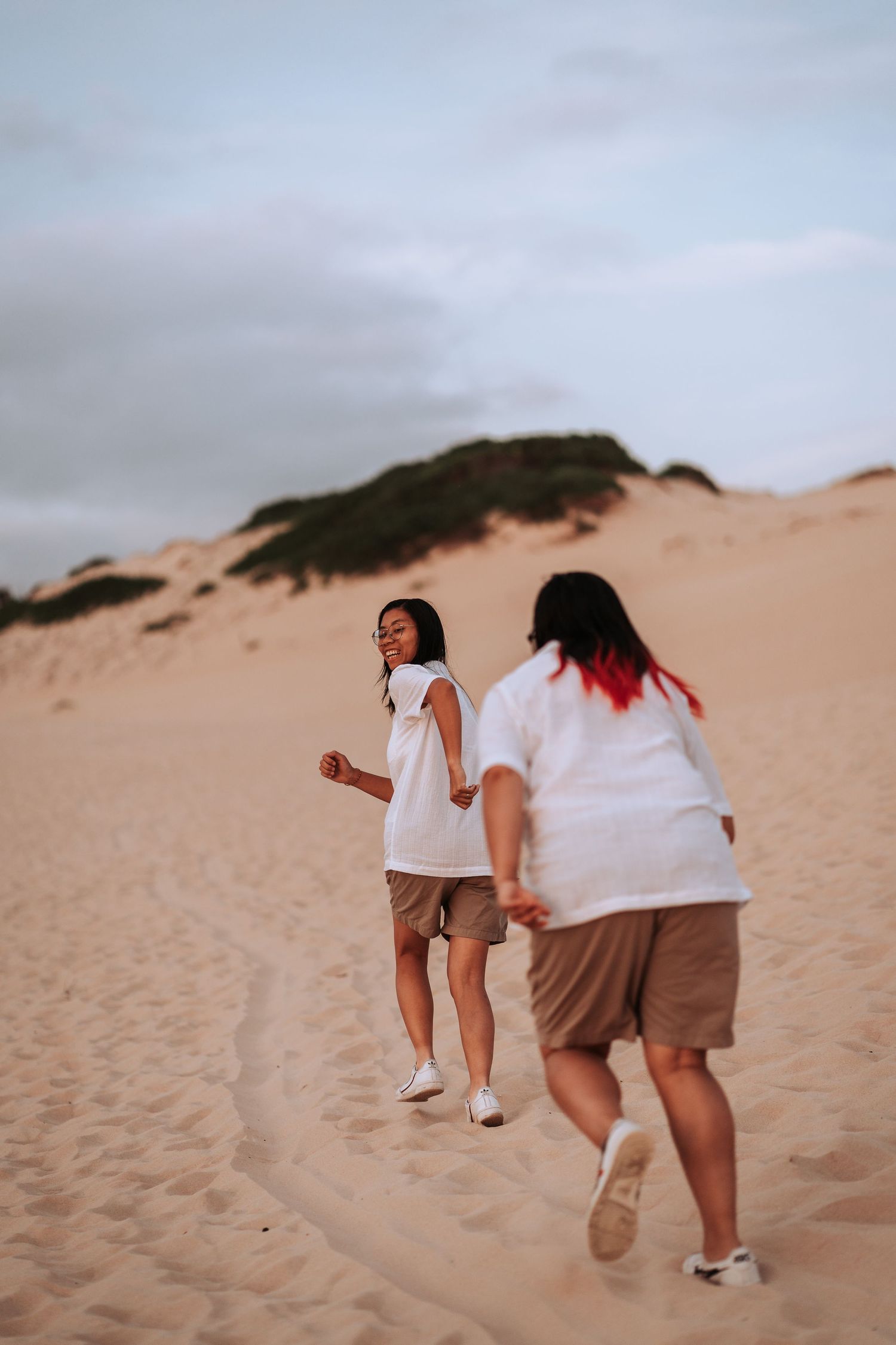 Two people walking together on sandy dunes at dusk wearing white tops and shorts against a cloudy sky.
