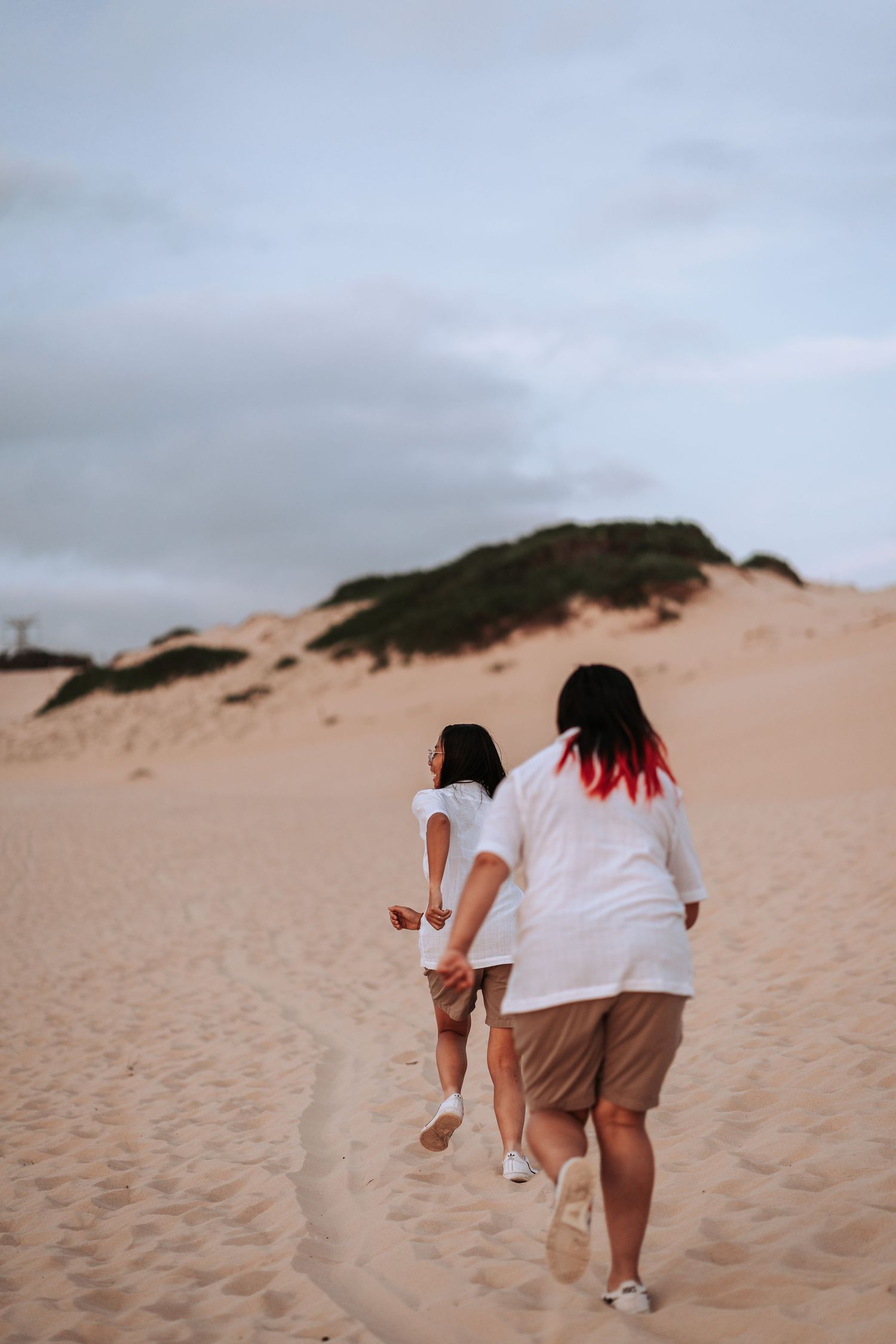 Two people in white shirts walking up sandy dunes against a cloudy sky at dusk.