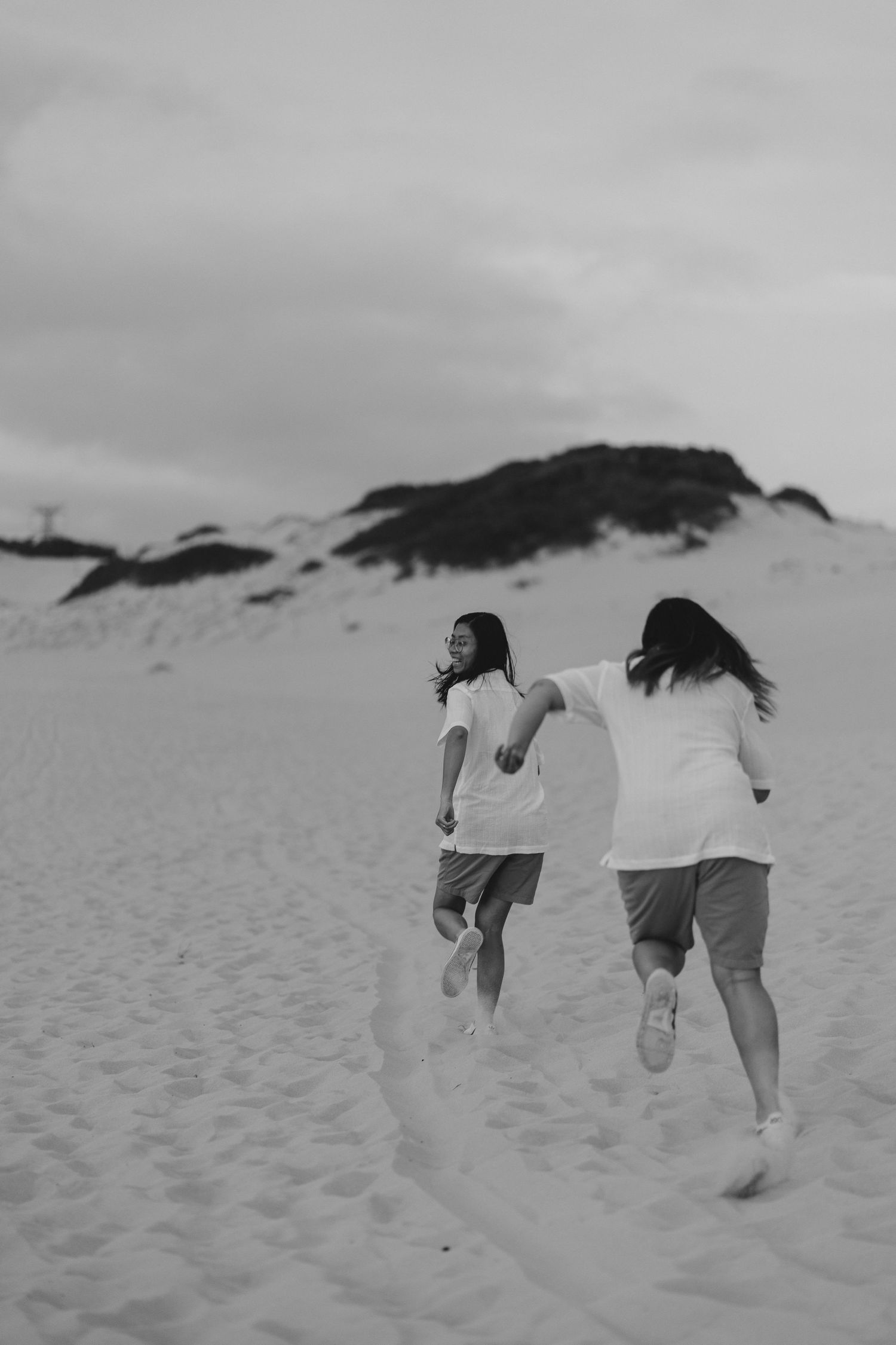 Two people in white shirts and shorts run joyfully across sandy dunes on a cloudy day, captured in black and white.