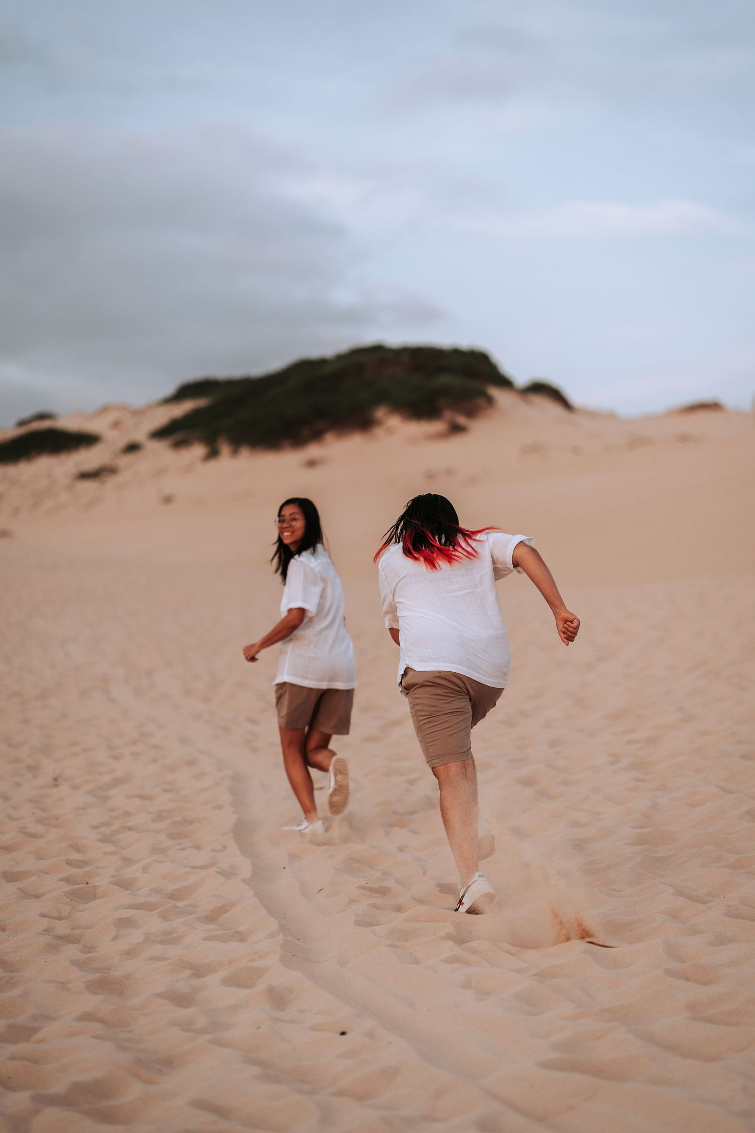 Two people in white shirts and brown shorts run playfully up sandy dunes under an overcast sky at sunset.