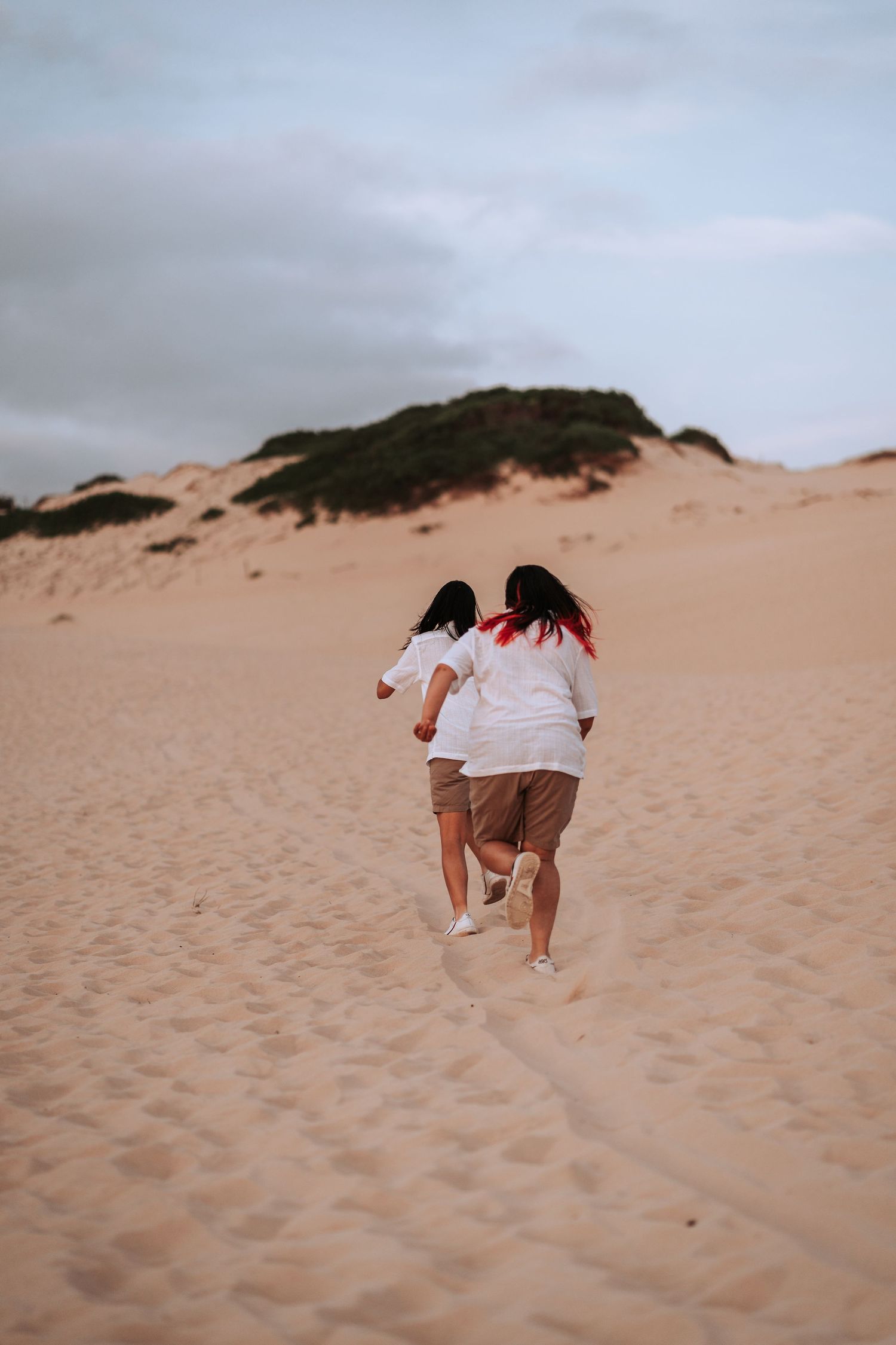 Two friends in white shirts run joyfully up a sandy dune under an overcast sky.