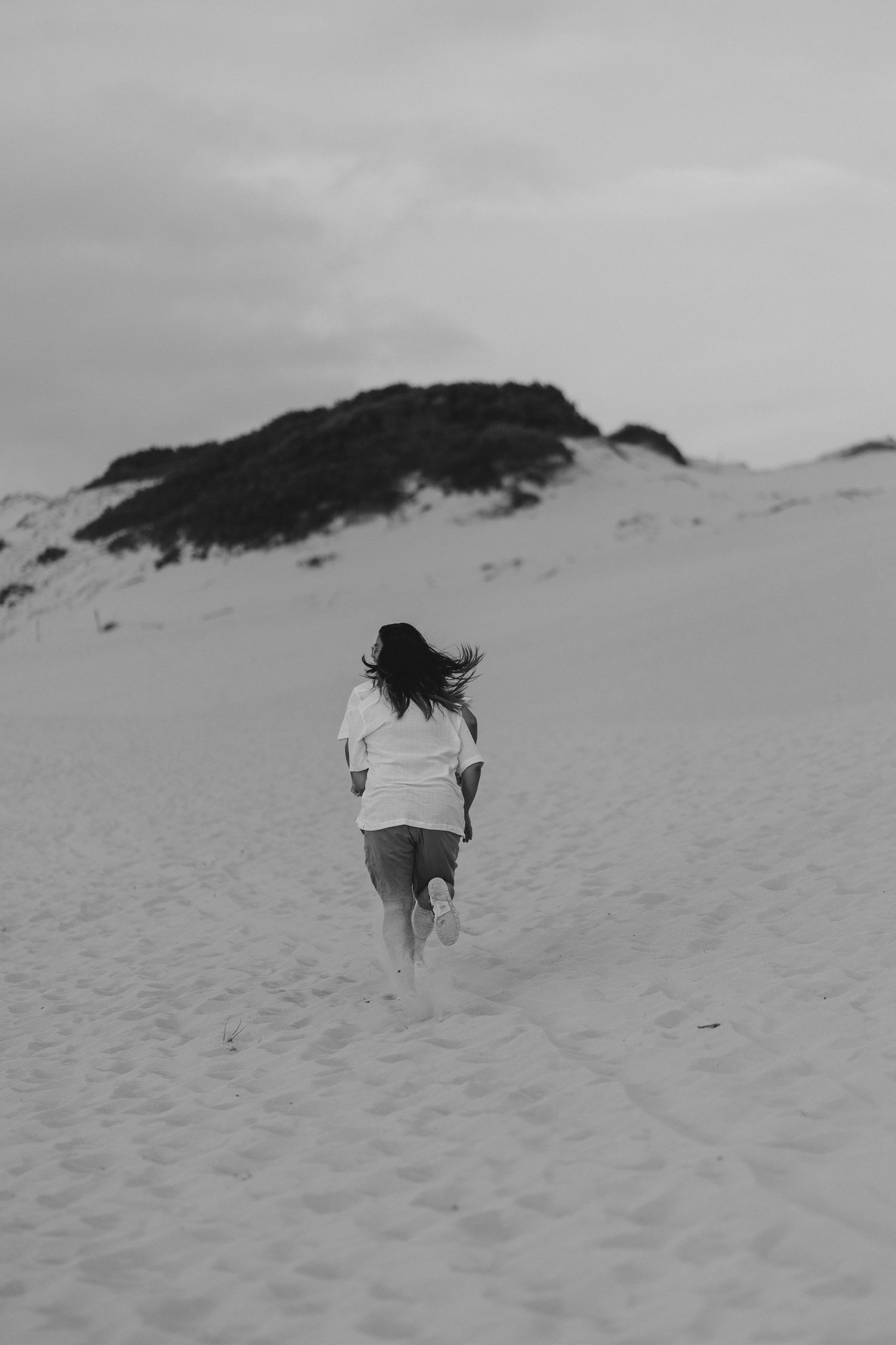 A lone figure in a white shirt walks up a snowy sand dune slope against an overcast sky in black and white.
