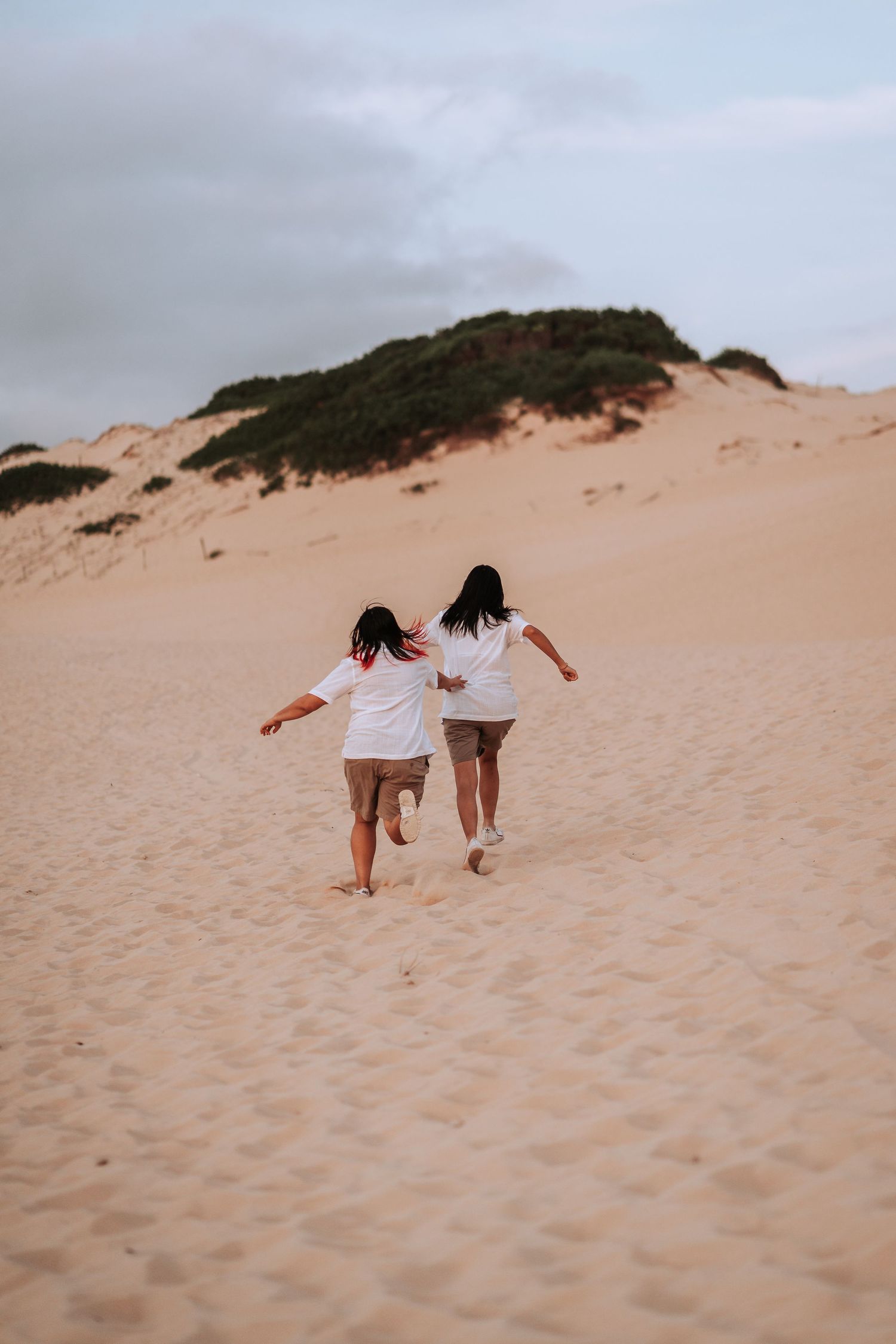 Two people in white outfits run together up a sandy dune under an overcast sky at sunset.