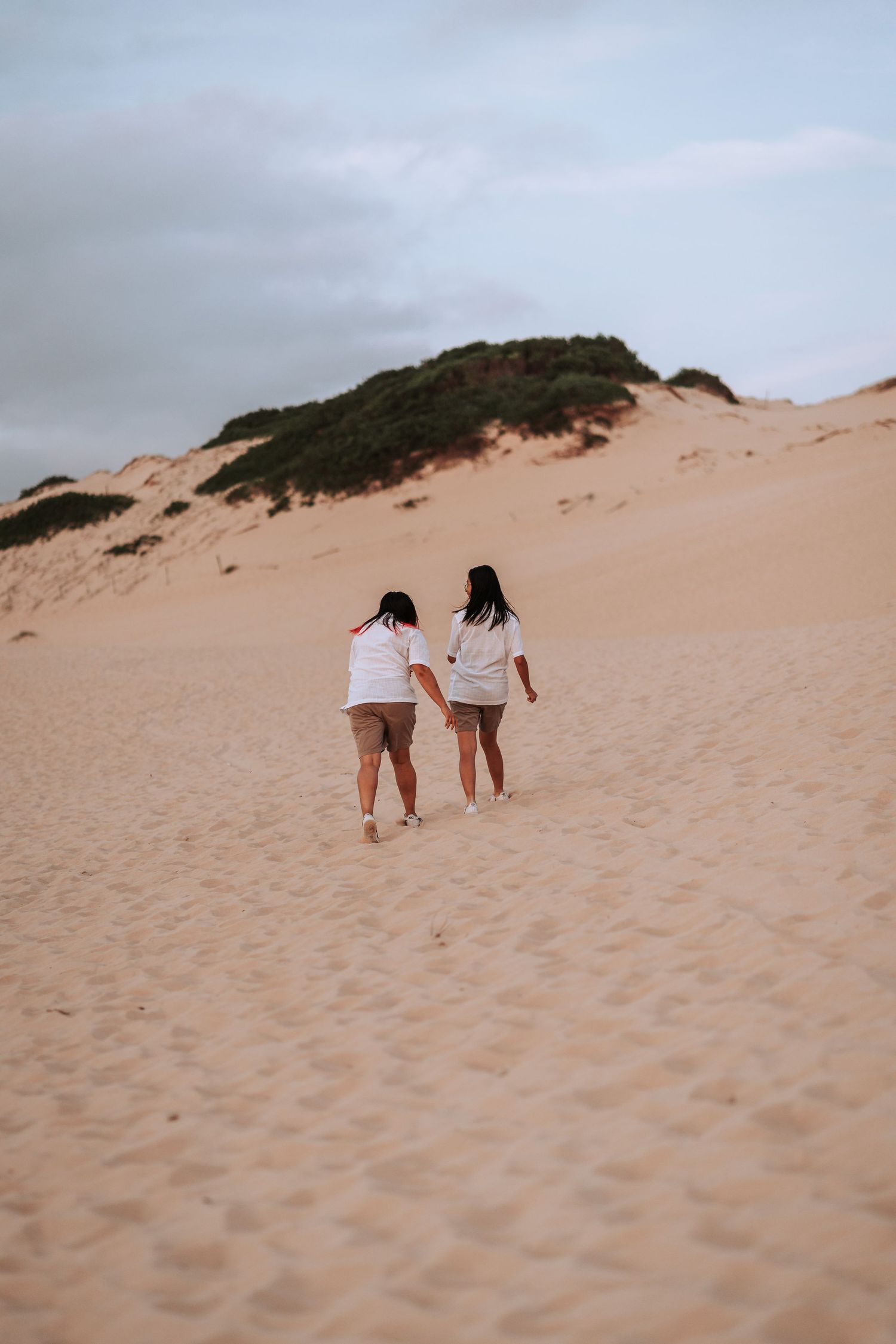 Two friends in matching outfits walk together up a sandy dune with vegetation in the background.