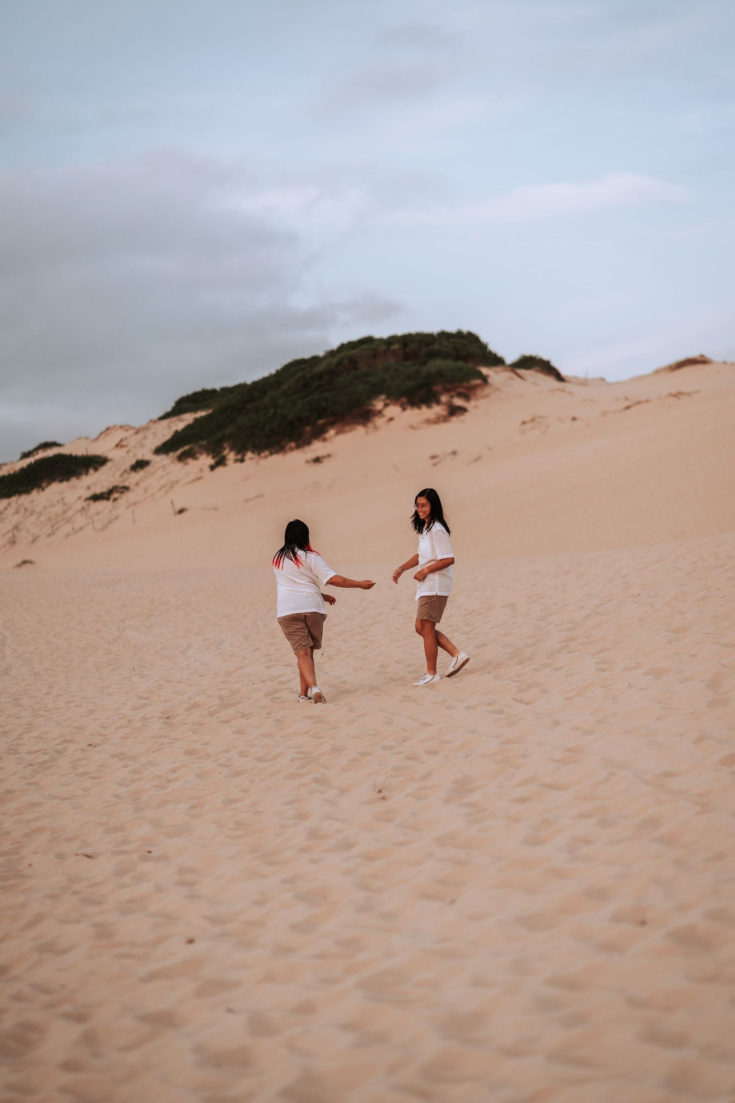 Two people playing and running together on a large sandy dune under an overcast sky.