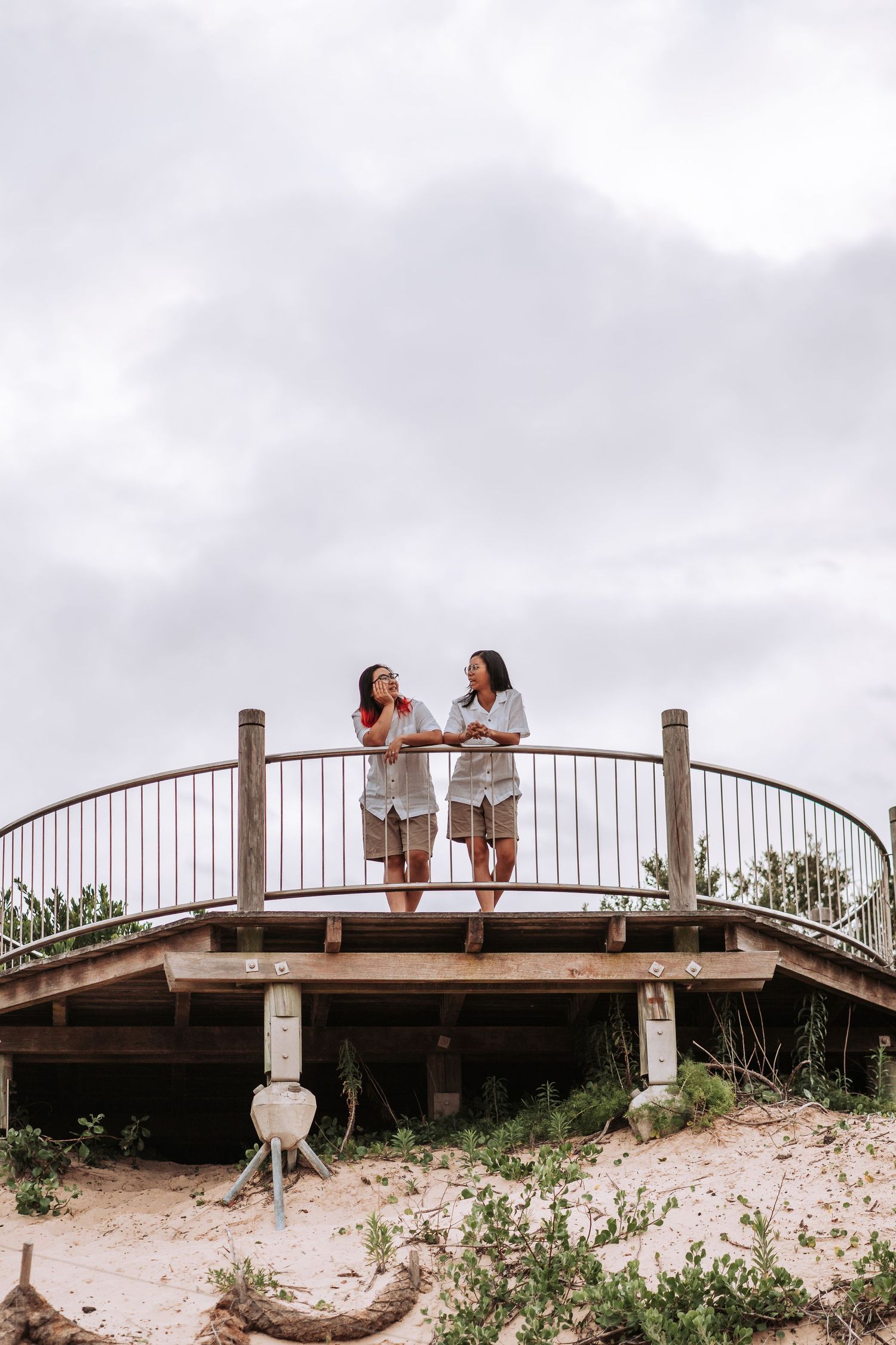Two people standing on a wooden footbridge overlooking a beach with cloudy skies in the background.