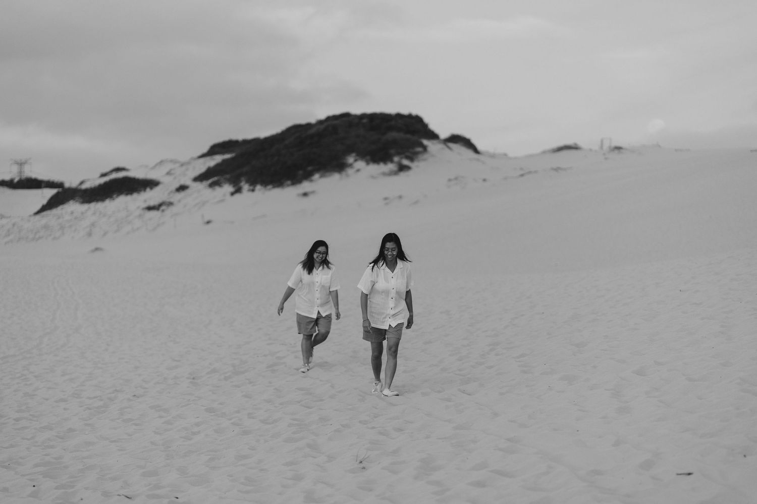 Two people in casual white t-shirts walk together on sand dunes in a black and white landscape.
